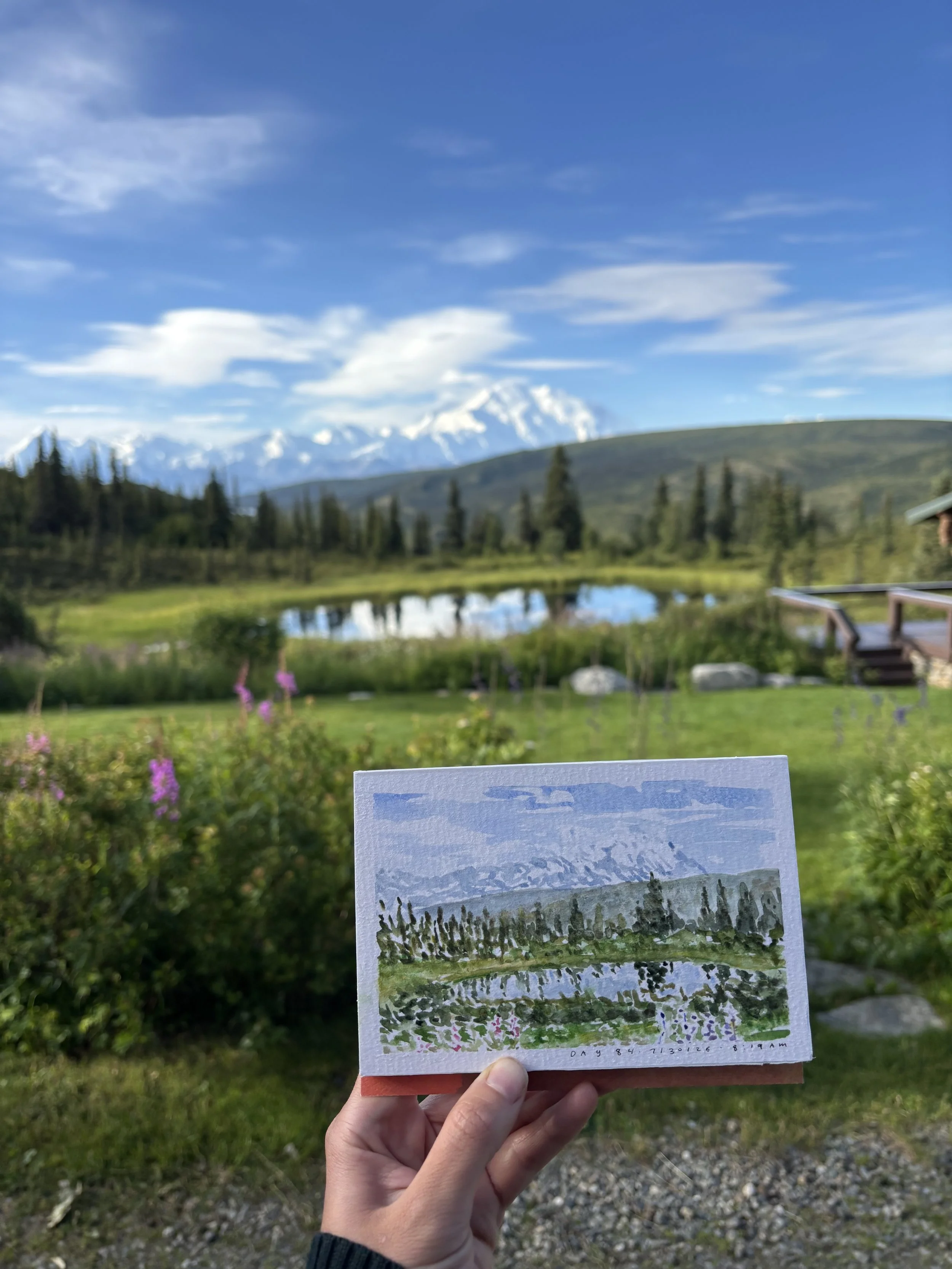 Hand holding a postcard with landscape painting, in front of a scenic view of mountains, trees, a lake, and a blue sky with some clouds.