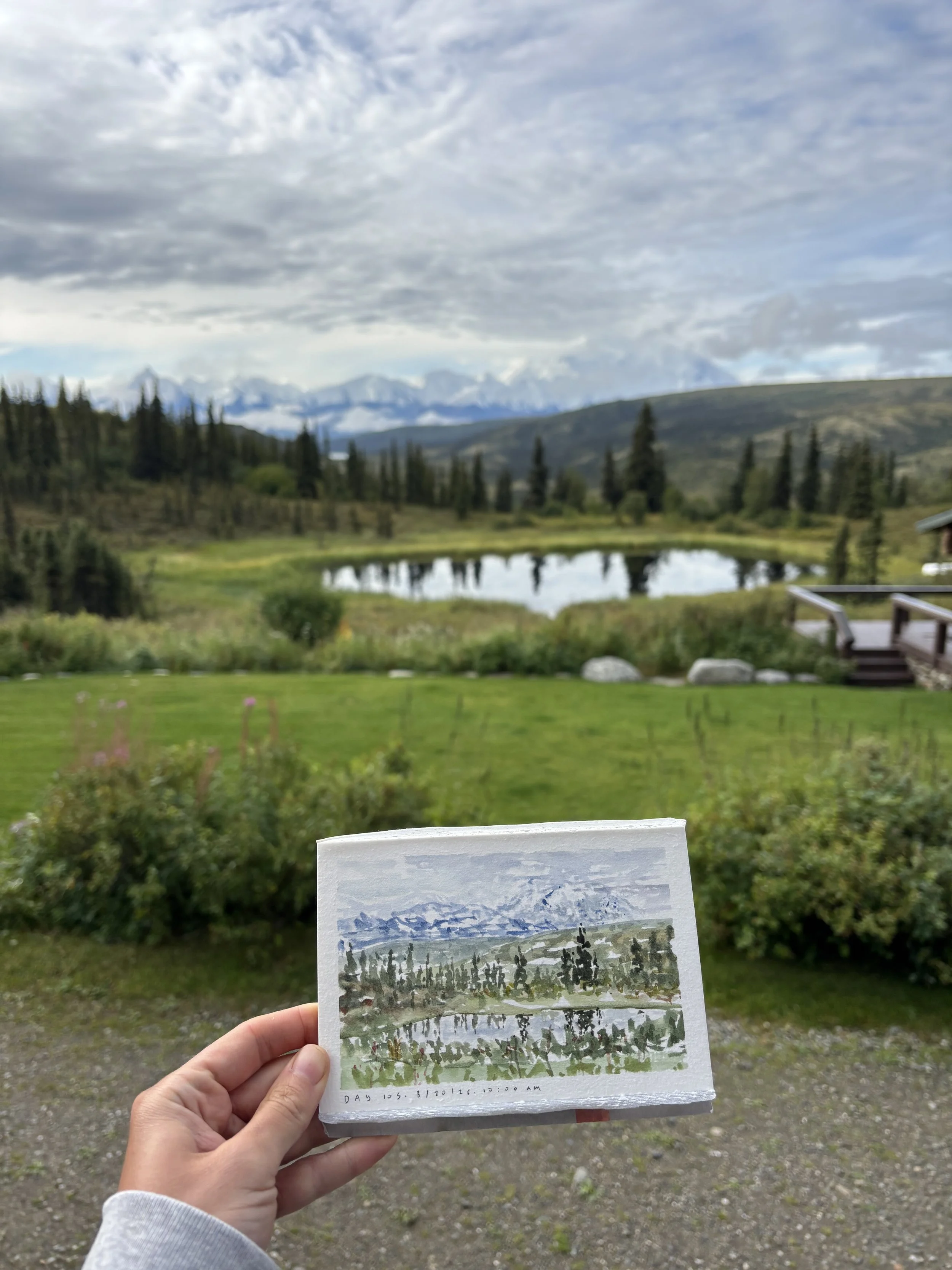 A person holding a small art print of a mountain landscape scene with trees in front of a mountain lake, with the actual landscape in the background including a green grassy area, trees, and distant snowy mountains under a cloudy sky.