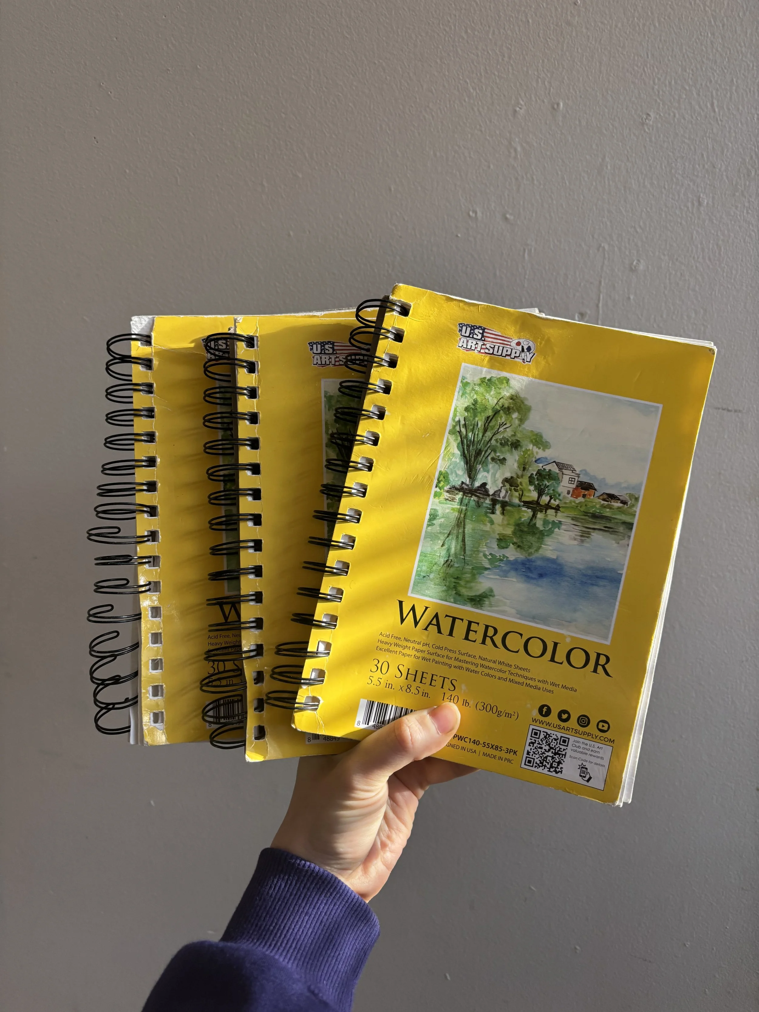 A hand holding a pack of 30 yellow watercolor paper sheets with a landscape painting of trees, houses, and water on the front cover.