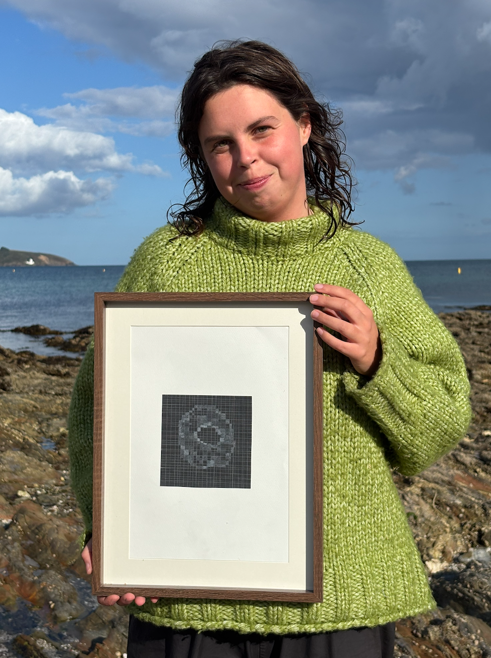A young woman with wavy brown hair, wearing a green knitted sweater, holding a framed digital artwork, standing on a rocky shoreline with the ocean and a cloudy sky in the background.