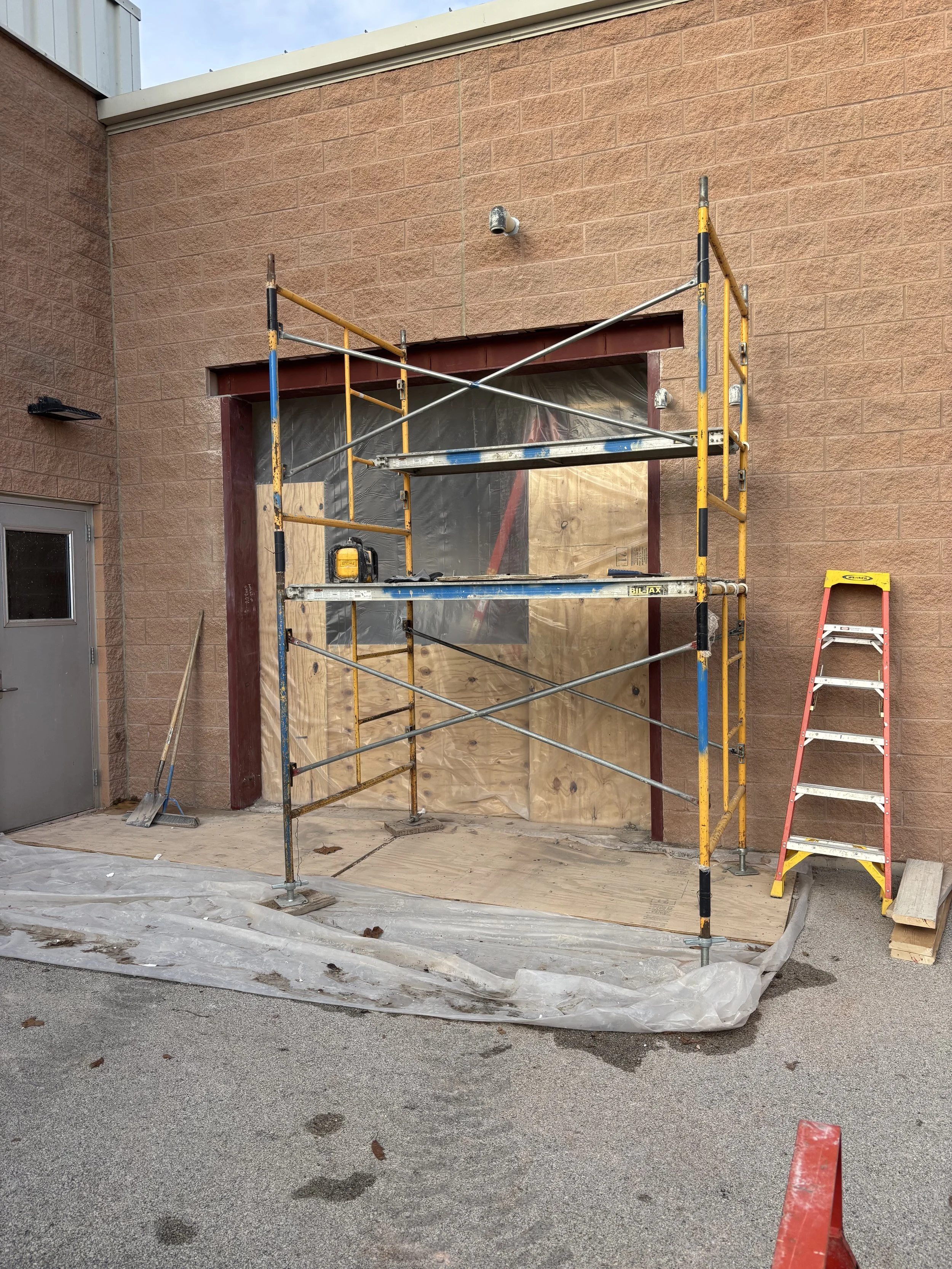 Construction scaffolding in front of a building with a large window opening covered with plastic sheeting, yellow and red ladders nearby, and construction tools and materials on the ground.