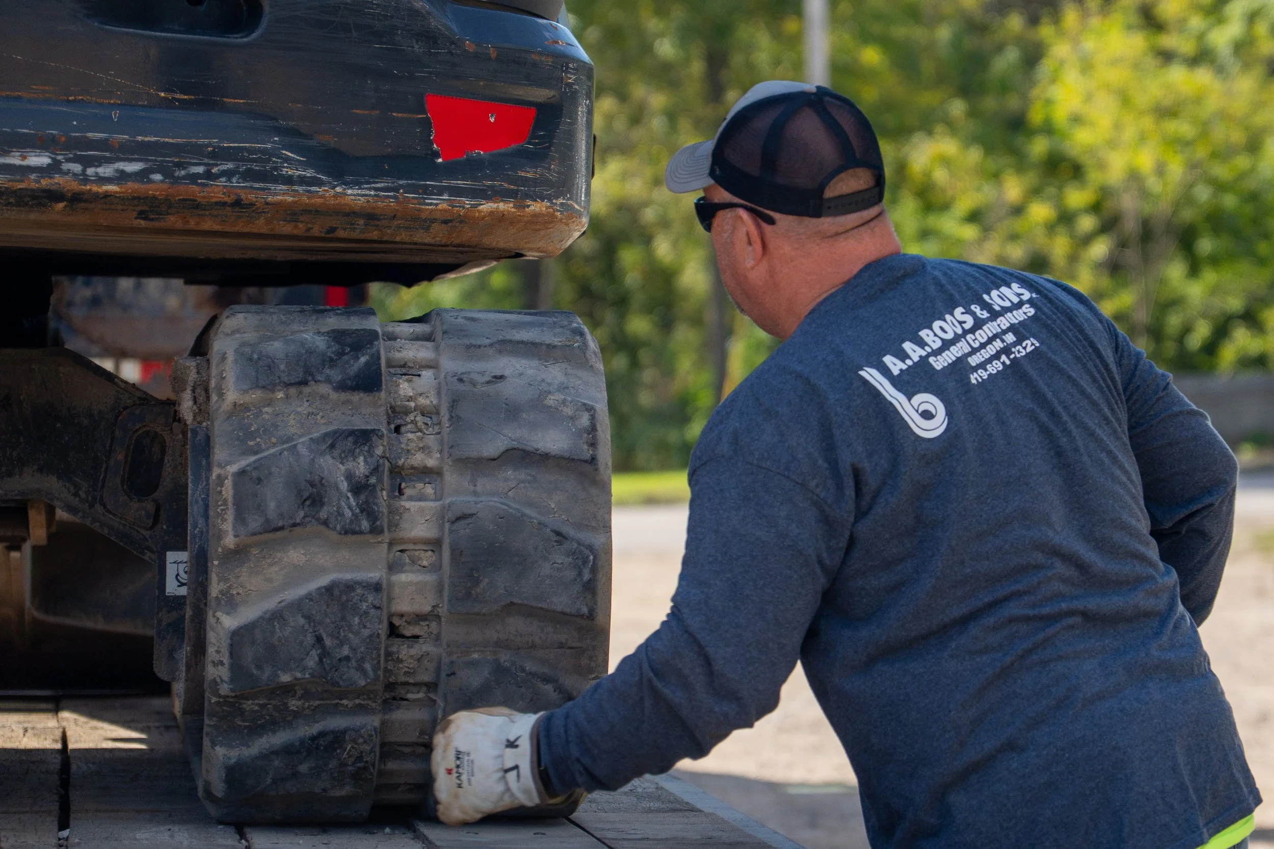 A person wearing a blue shirt, black cap, and sunglasses is working on a construction or machinery project outdoors, possibly repairing or maintaining equipment. The person's shirt has text on the back that reads 'A. A. BOIS & SON, General Contractors, Oregon, IL, phone number 419-691-1324'. The image shows part of a large, dirty, muddy piece of machinery or vehicle, with a red reflector and some rust, set against a background of green trees.
