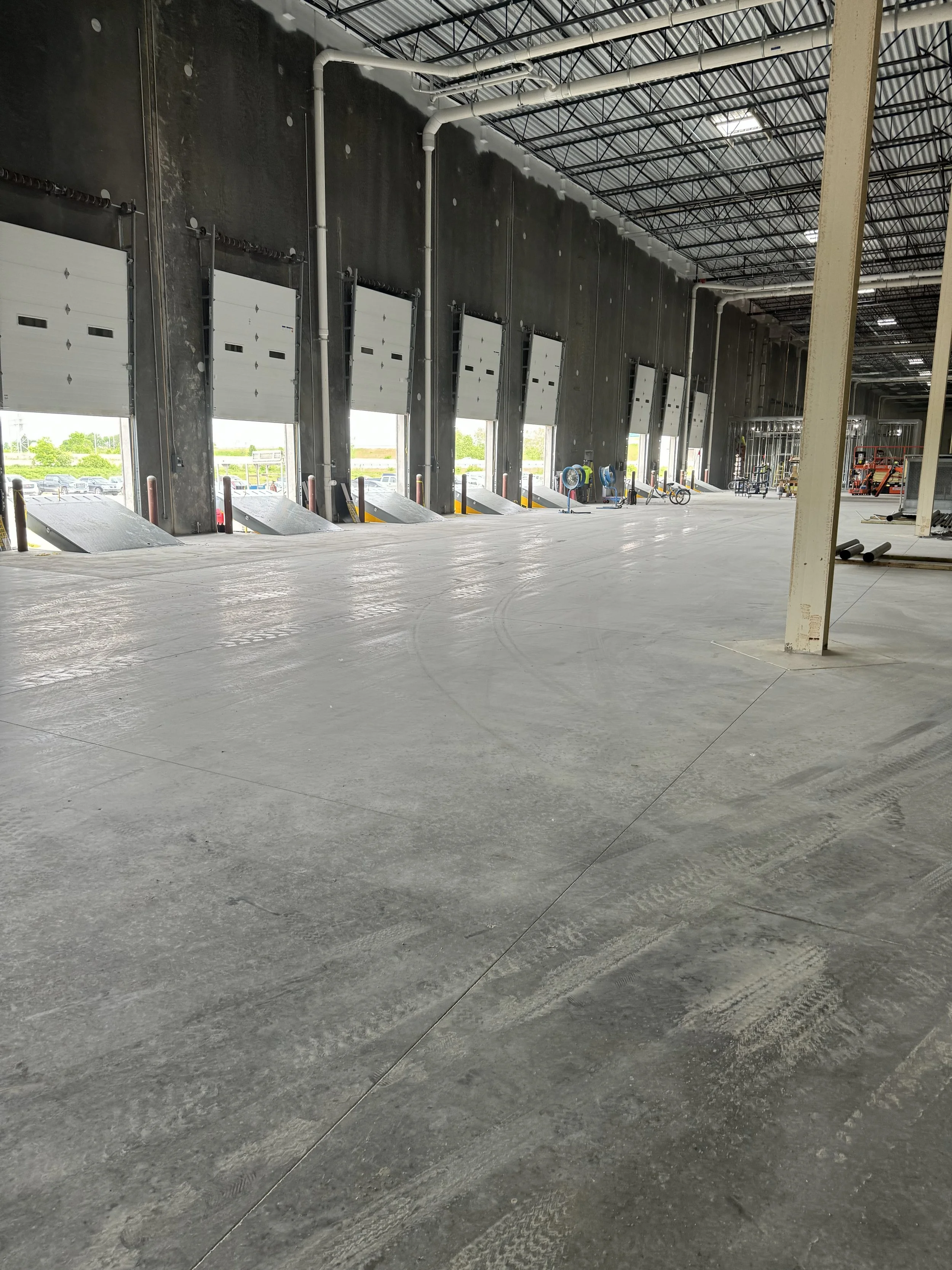 Inside view of a large warehouse or distribution center under construction, with multiple white garage doors, concrete floors, and industrial ceiling with pipes and skylights.