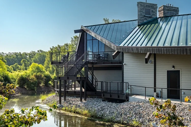 A house with a metal roof and outdoor staircase beside a body of water, surrounded by greenery.