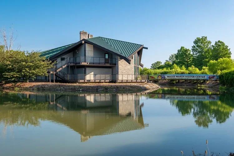 A modern visitor center building with a sloped green roof, situated next to a pond that reflects the building and surrounding trees. A sign reads 'Magee Marsh Visitor Center'.