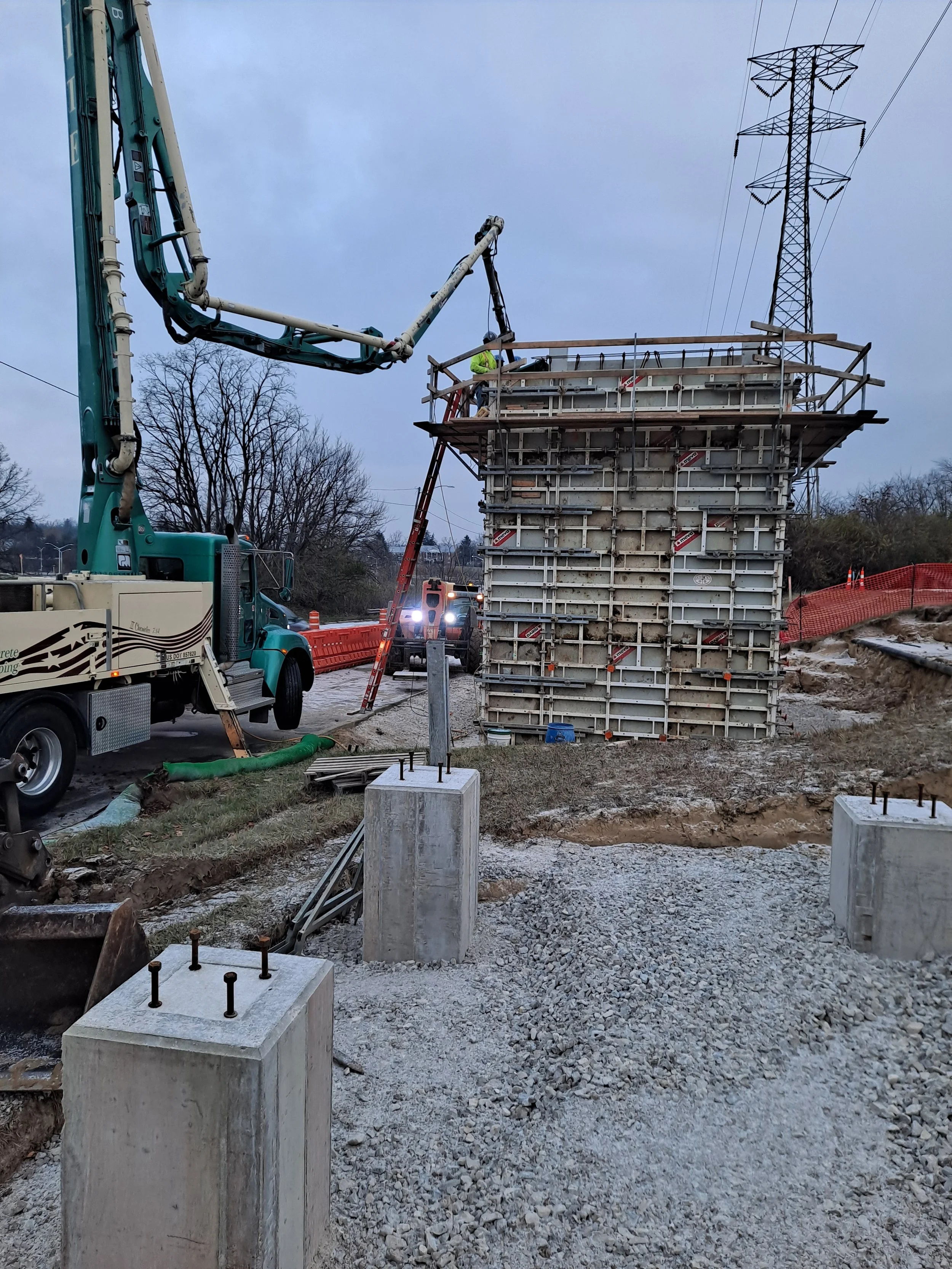 Construction site with concrete forms, scaffolding, and a crane. Worker in high-visibility vest working on the structure, with power lines and a transmission tower in the background.