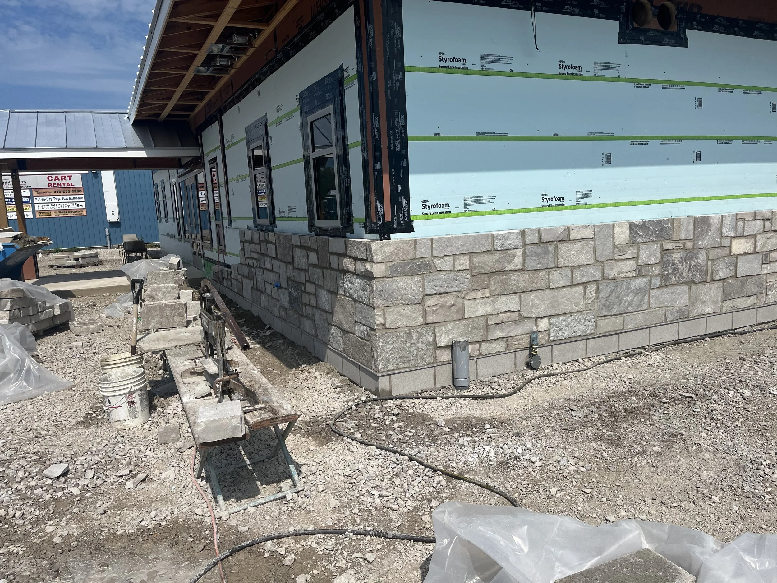 Construction site showing partially built house with a stone foundation, blue and white exterior walls, and windows. Building materials and tools are scattered around, and the sky is clear.
