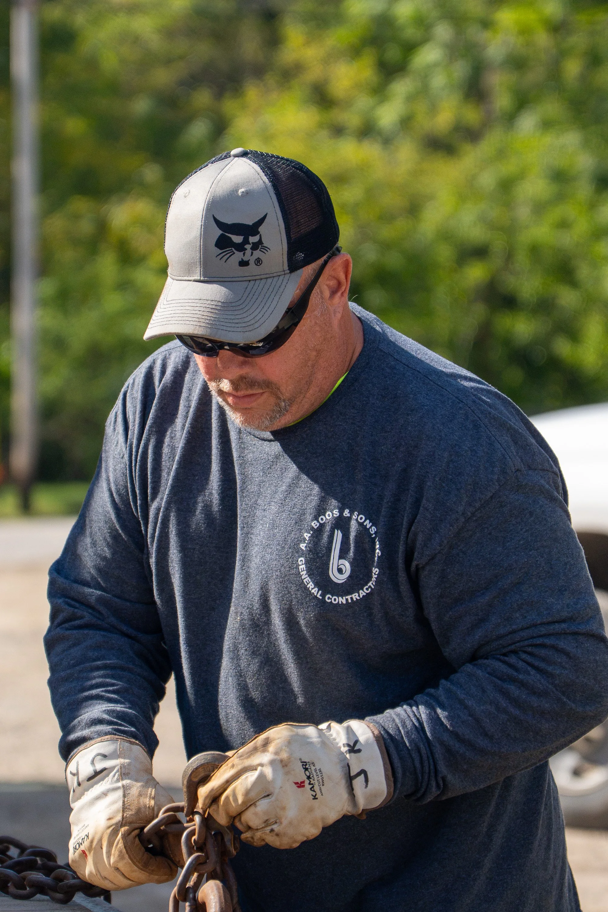 A man wearing a gray and black baseball cap with a black panther logo, sunglasses, a gray shirt, and work gloves is handling a chain outdoors with trees blurred in the background.
