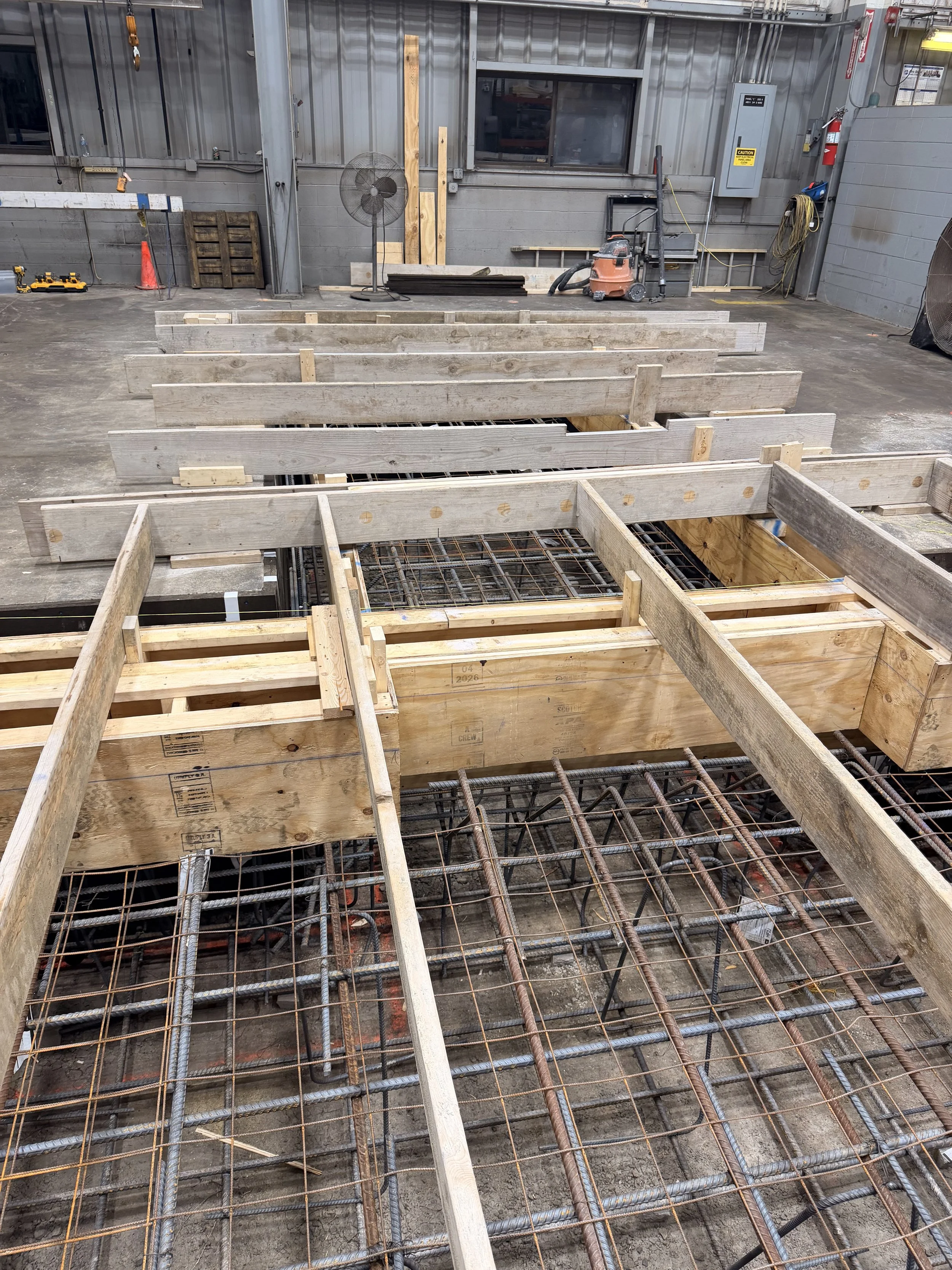Construction site with wooden formwork and steel rebar on the ground, inside a workshop with metal walls, tools, a fan, and construction equipment.