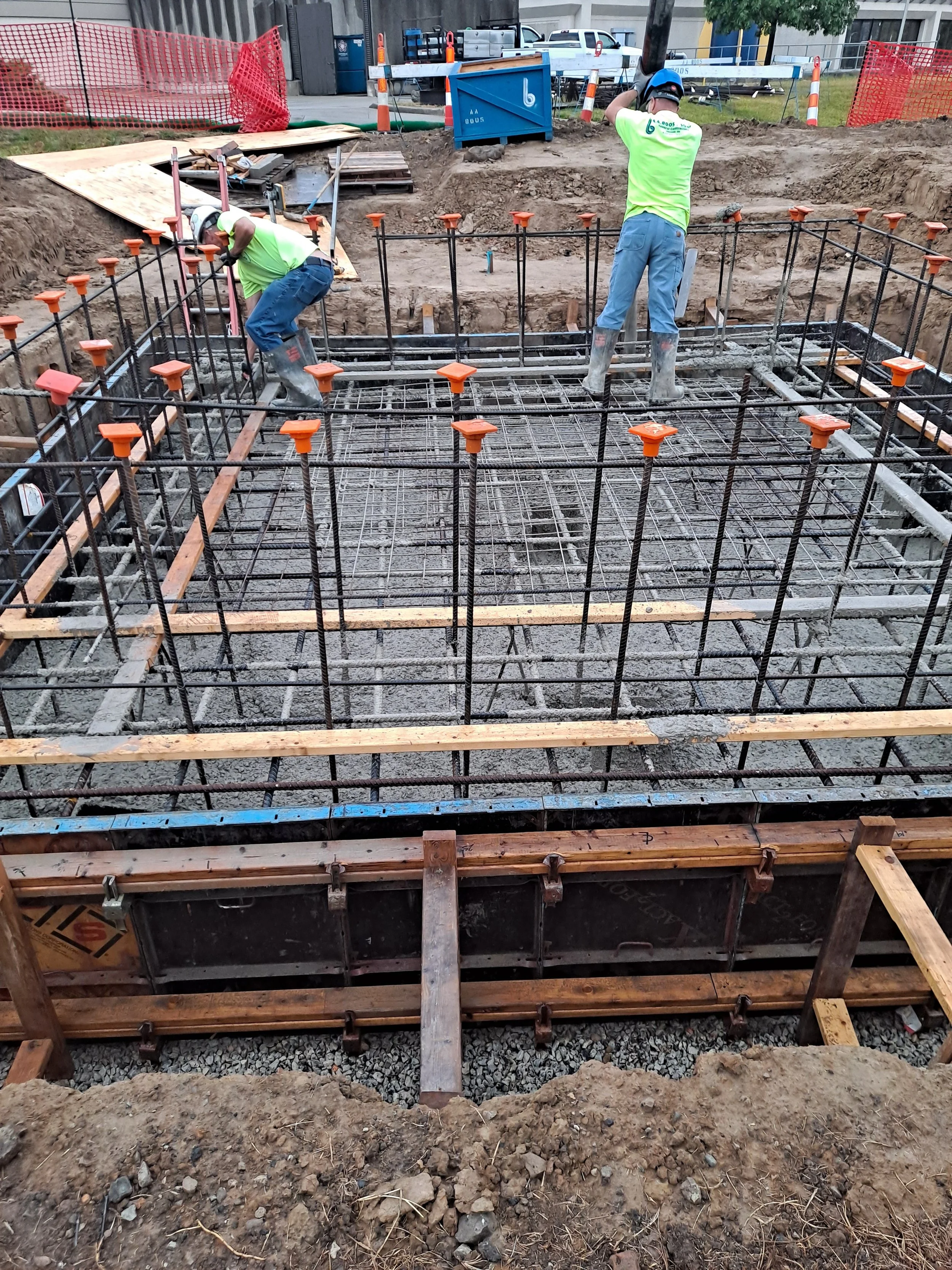 Construction site with two workers wearing high-visibility shirts and helmets, working on pouring concrete into a foundation with metal rebar reinforcement and wooden support beams. There are construction tools, barriers, and a truck in the background.