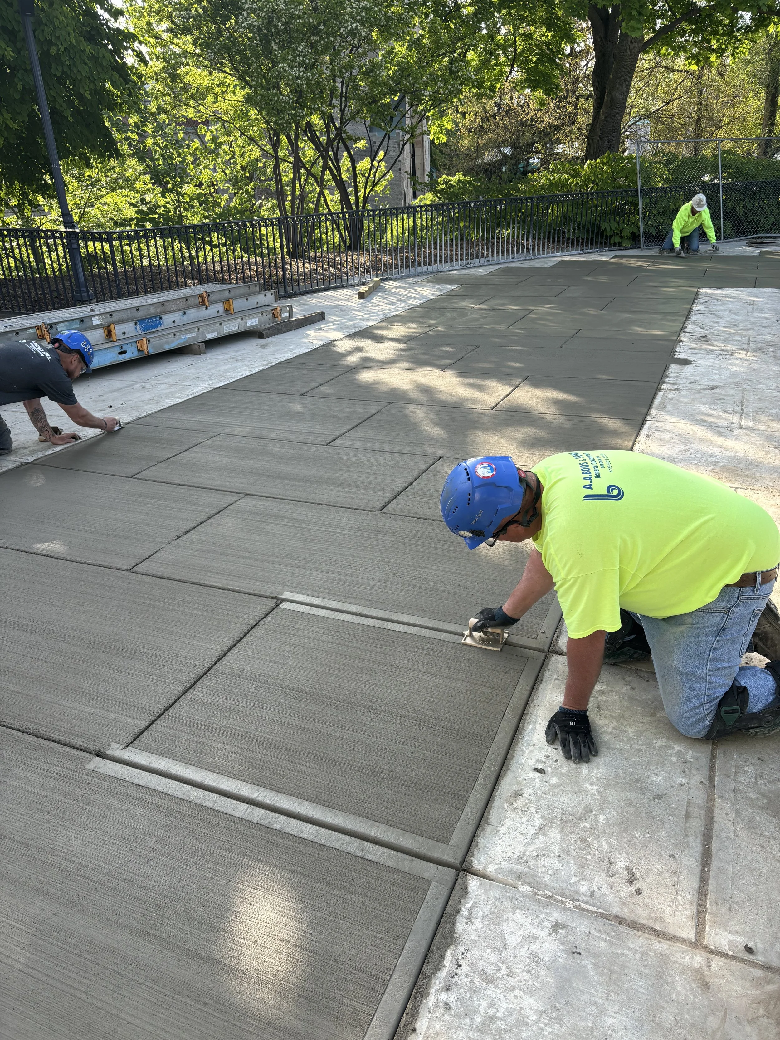 Construction workers installing or repairing large paving slabs on a sidewalk surrounded by trees and a fence, with one worker kneeling and another crouching nearby.