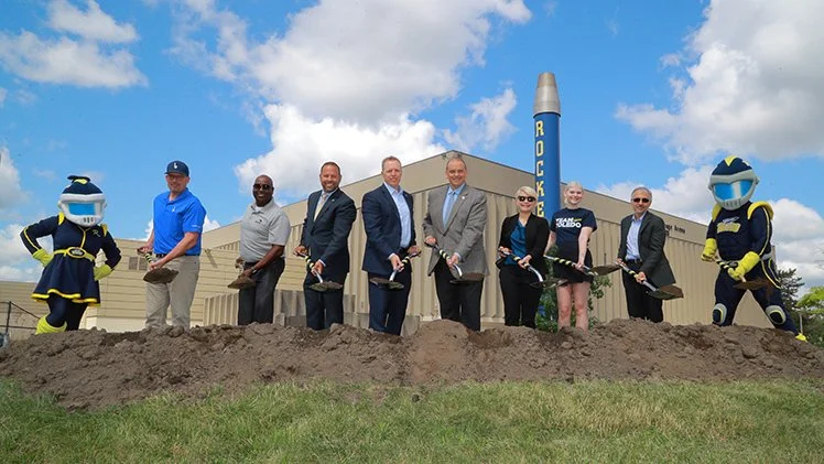 Group of people, including two mascots, participating in a groundbreaking ceremony with shovels in front of a building and a sign that says 'ROCKET' on a large chimney.