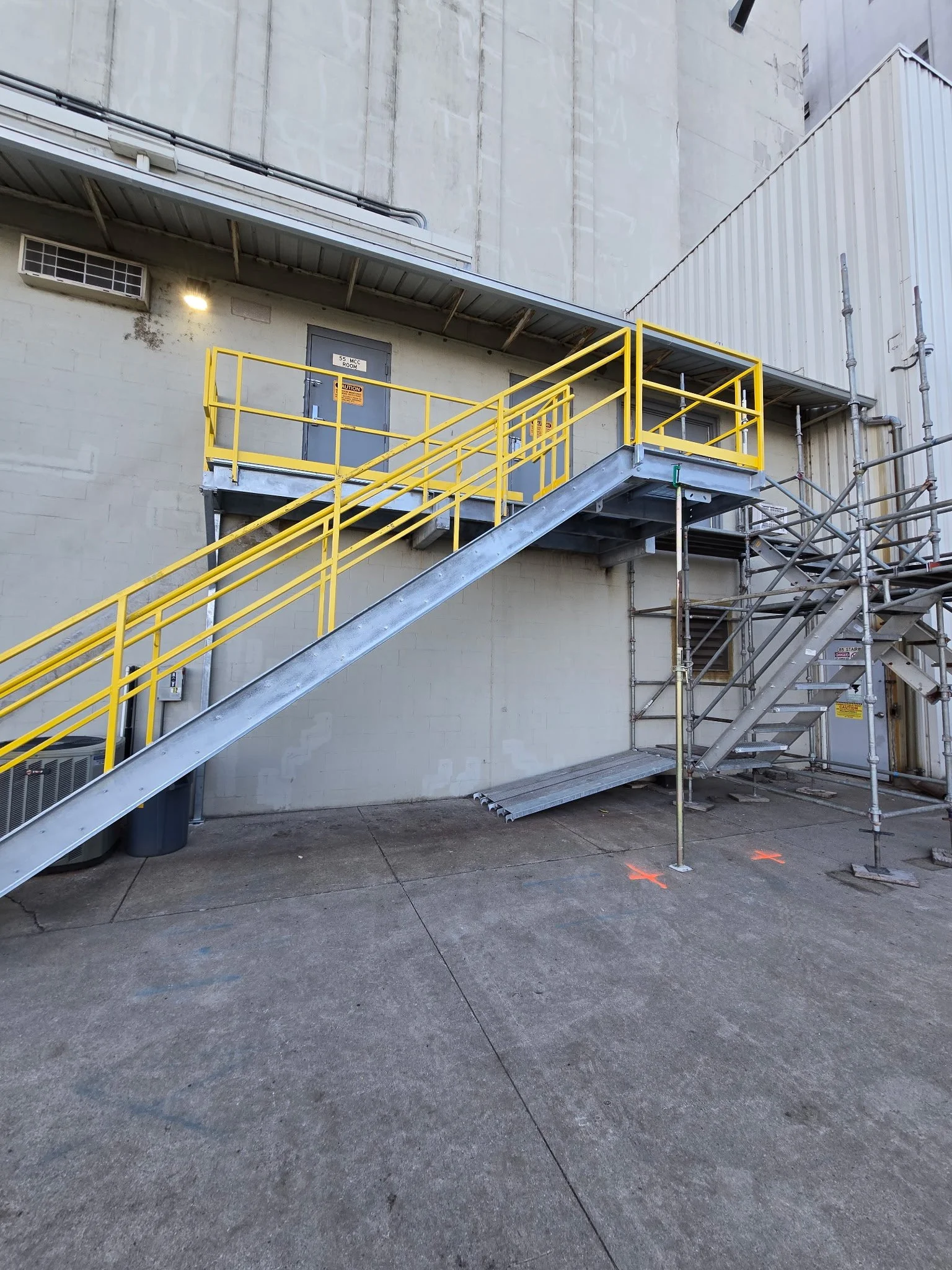 Exterior city scene with metal staircase, yellow railings, concrete sidewalk, and scaffolding on the right side. The building has a beige wall, a door marked '55 MCC ROOM,' a vent, and a light. The ground has orange markings.