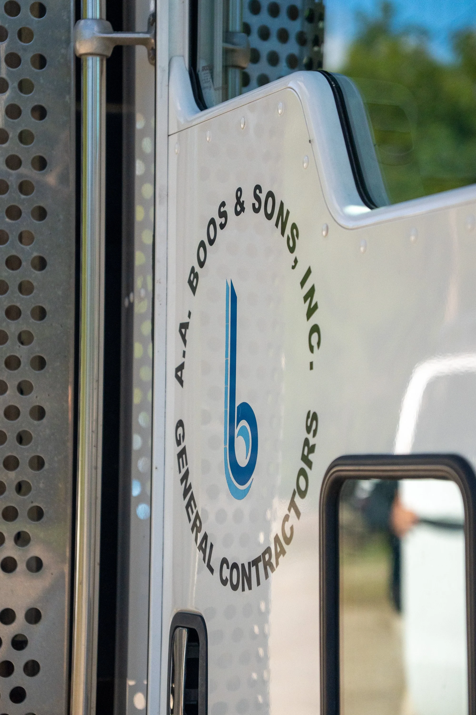A vehicular license plate with a circular sticker that has the logo 'G' in blue and the words 'Alabama, Booth, Inc., Professional Contracting' written around the circle, mounted on a metal frame with a perforated structure.
