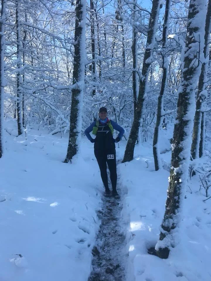 A person standing on a snow-covered trail in a winter forest with snow-laden trees in the background. The individual is wearing dark clothing, a blue jacket, and a green backpack, with hands on hips, and appears to be participating in a winter outdoor activity.
