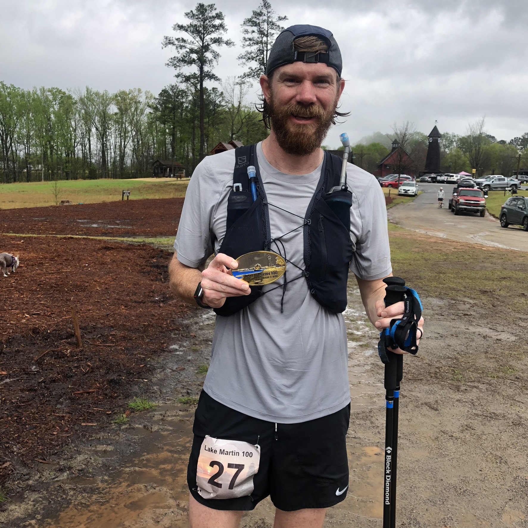 Man with a beard and cap holding a medal, wearing running gear and a race bib number 27, standing outdoors on a muddy trail after a race.