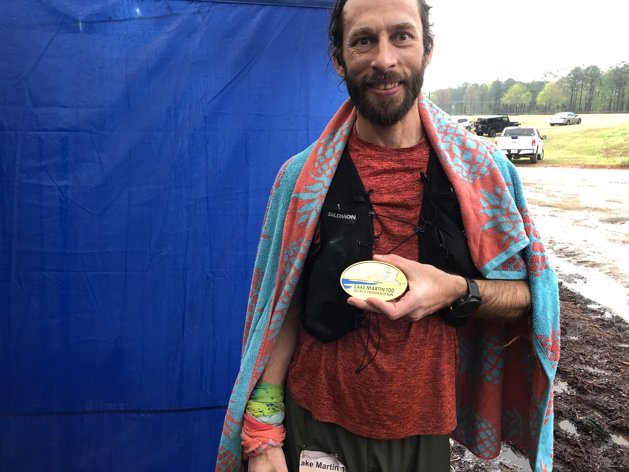 Man outdoors after a race, holding a medal that reads 'Lake Martin 100'. He is smiling, wrapped in a colorful towel, and wearing a red shirt, black vest, and sports watch. In the background, there are cars on a gravel area and a grassy field with trees.