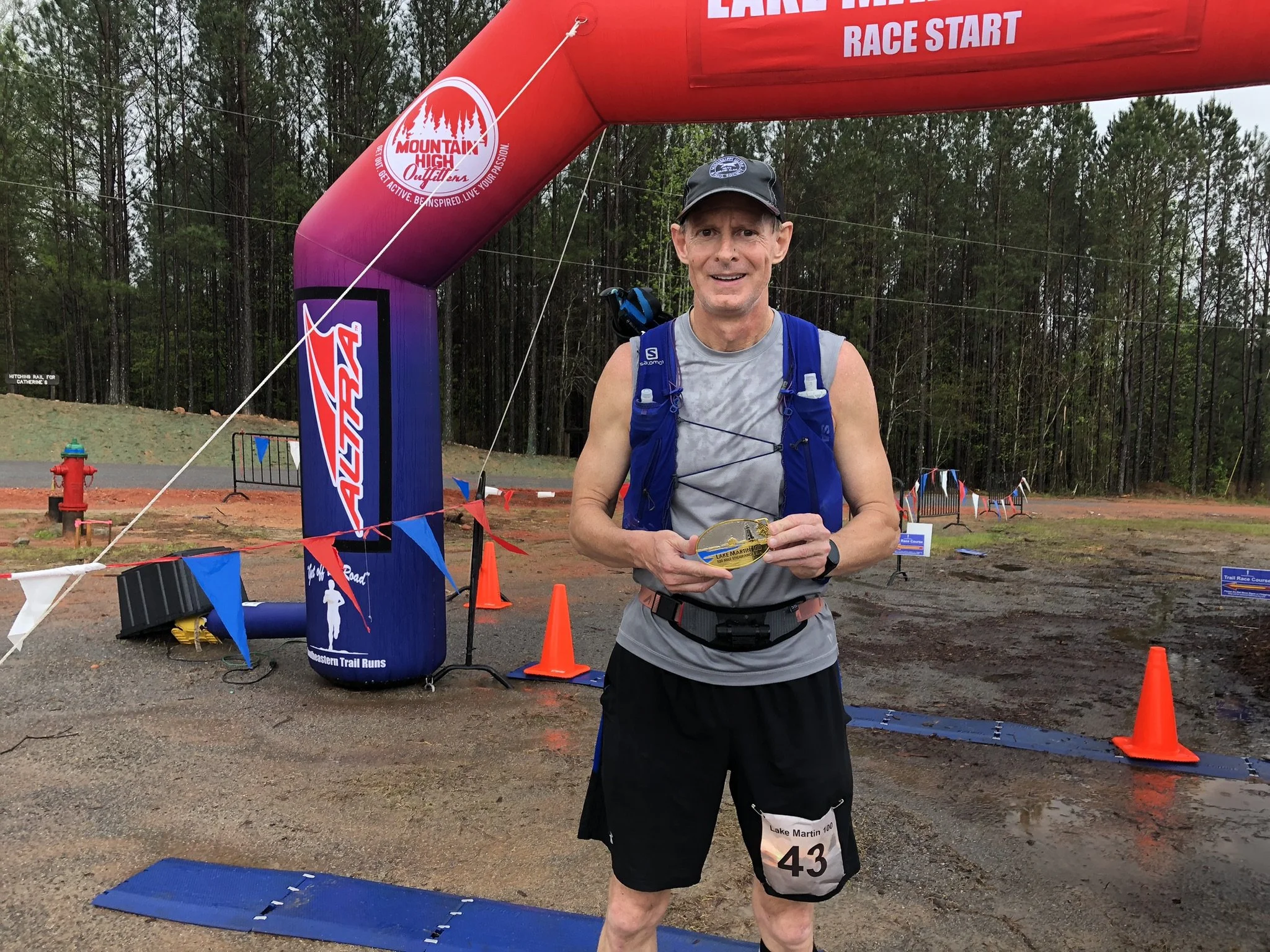A man in running attire with race bib number 43 stands in front of a finish line at an outdoor trail race, holding a medal, with an inflatable arch and trees in the background.