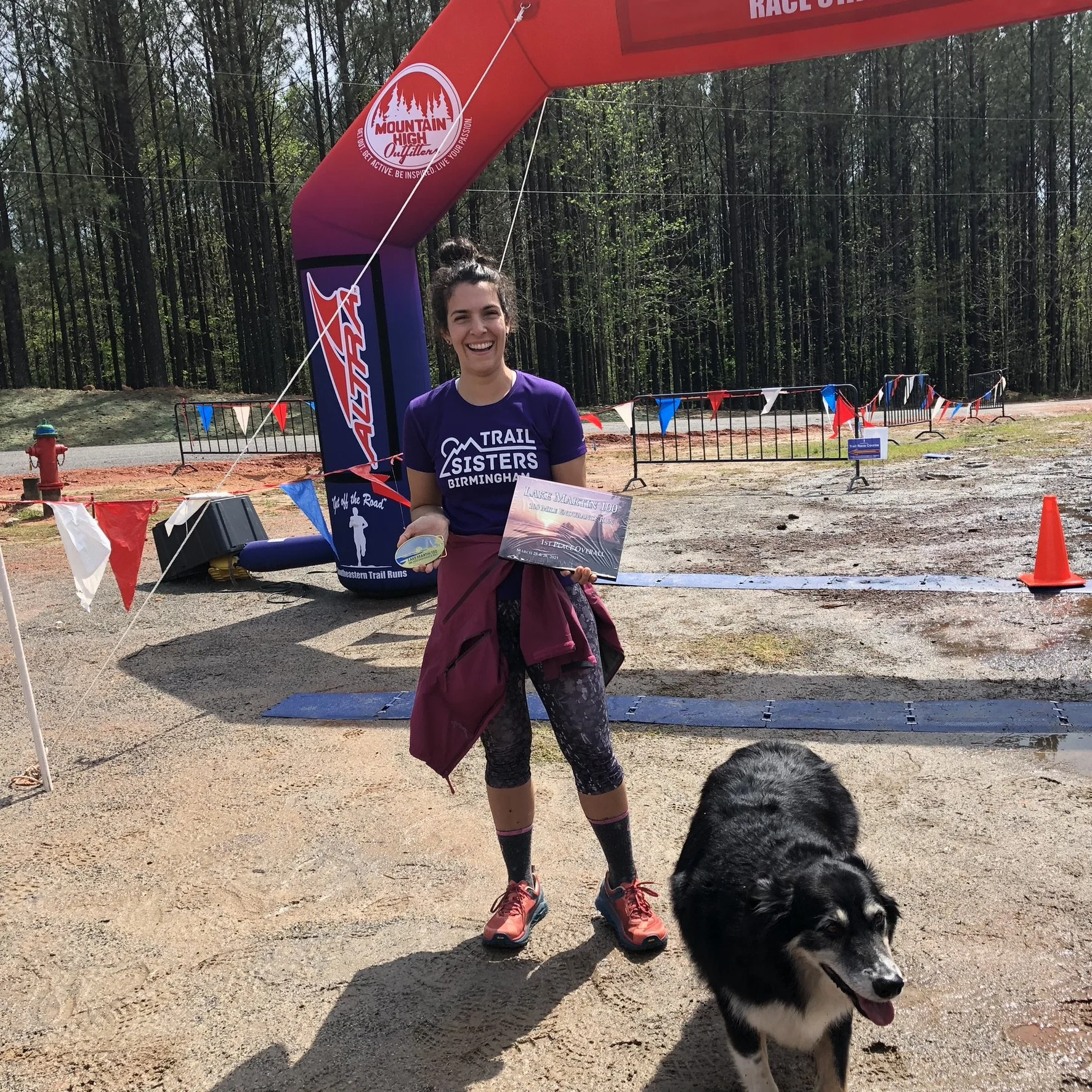 A woman in athletic gear holding a medal and a certificate, smiling in front of a large red finish line arch at a trail running event, with her dog standing nearby. Trees are in the background.