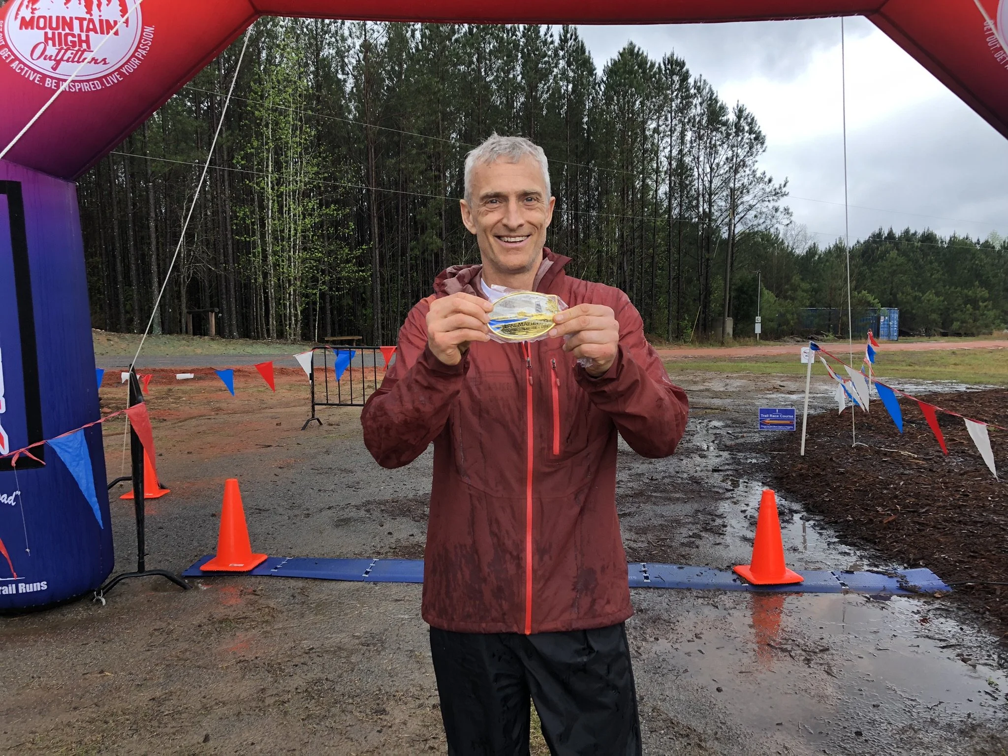 A man in a red jacket holding a medal at an outdoor event with a finish line arch and orange cones amid a muddy, rainy environment, with trees in the background.