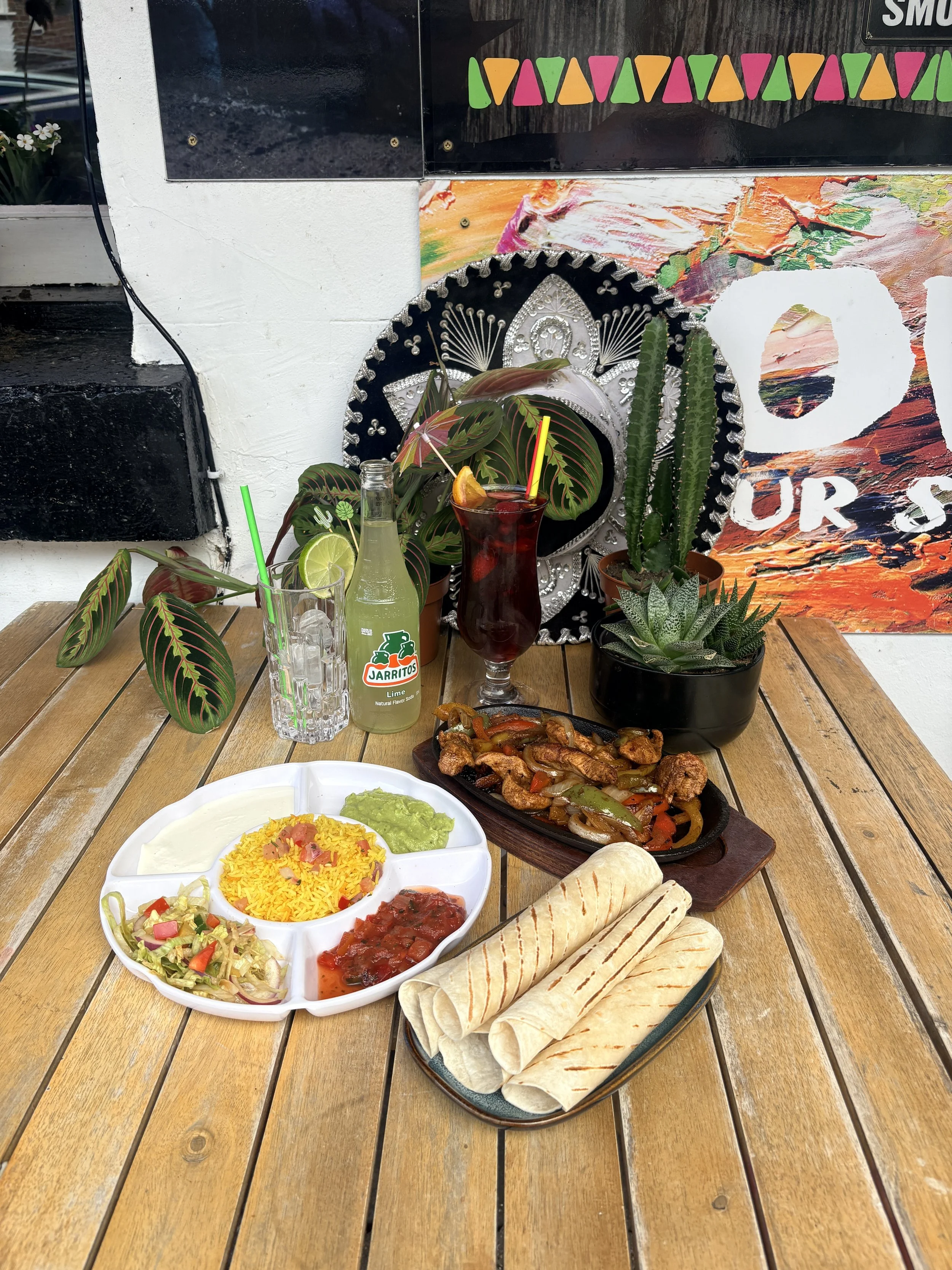 A wooden table holding a Mexican meal including a white divided plate with rice, guacamole, pico de gallo, and salsa, a black oval platter with cooked meat and vegetables, a stack of grilled flour tortillas, and two beverages—one lime soda and one hibiscus drink with a lemon wedge and striped straw. In the background, there are various plants and decorative items, including a cactus, a sombrero, and colorful posters on the wall.