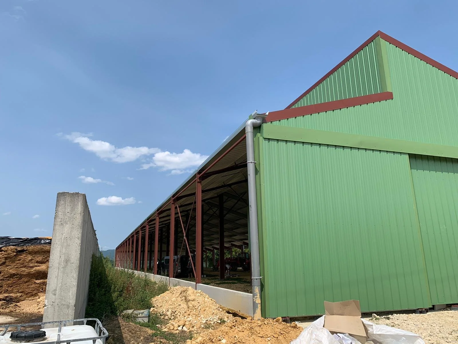 Ein grünes Lagergebäude unter Bau, mit einer offenen Seite, Baufahrzeugen und Baustellenmaterial im Vordergrund, blauer Himmel mit wenigen Wolken im Hintergrund.