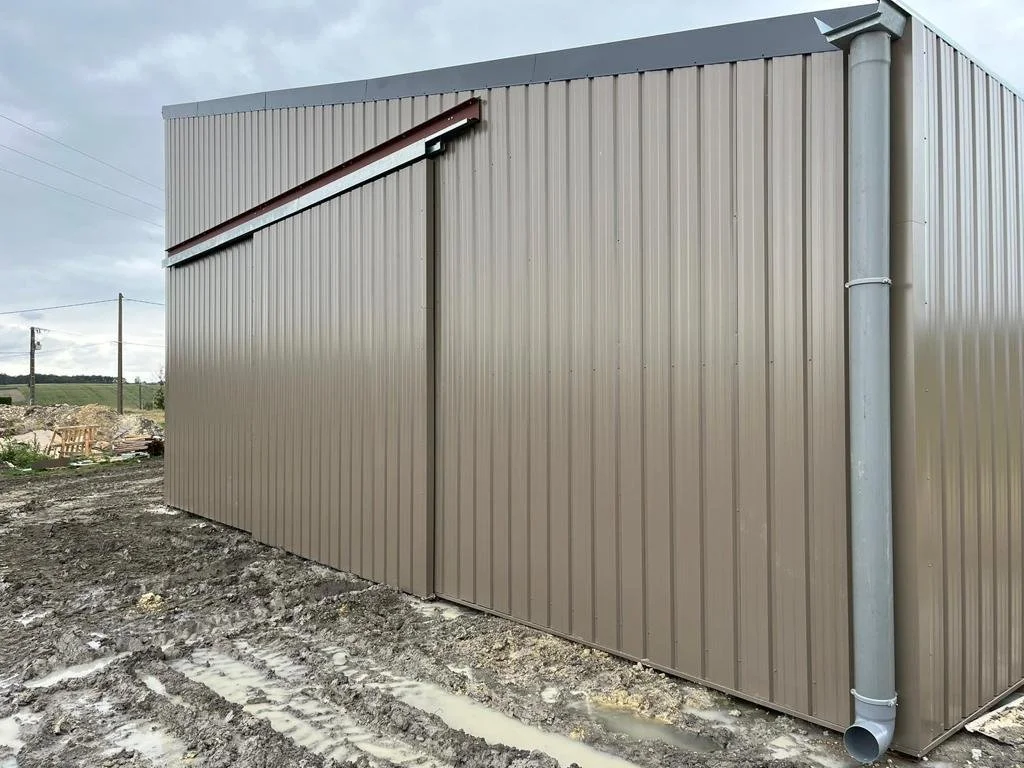 Ein Gebäude mit metallischer Fassade, seitlich ein Fallrohr und eine Regenrinne, im Hintergrund eine Baustelle und Stromleitungen bei bewölktem Himmel.
