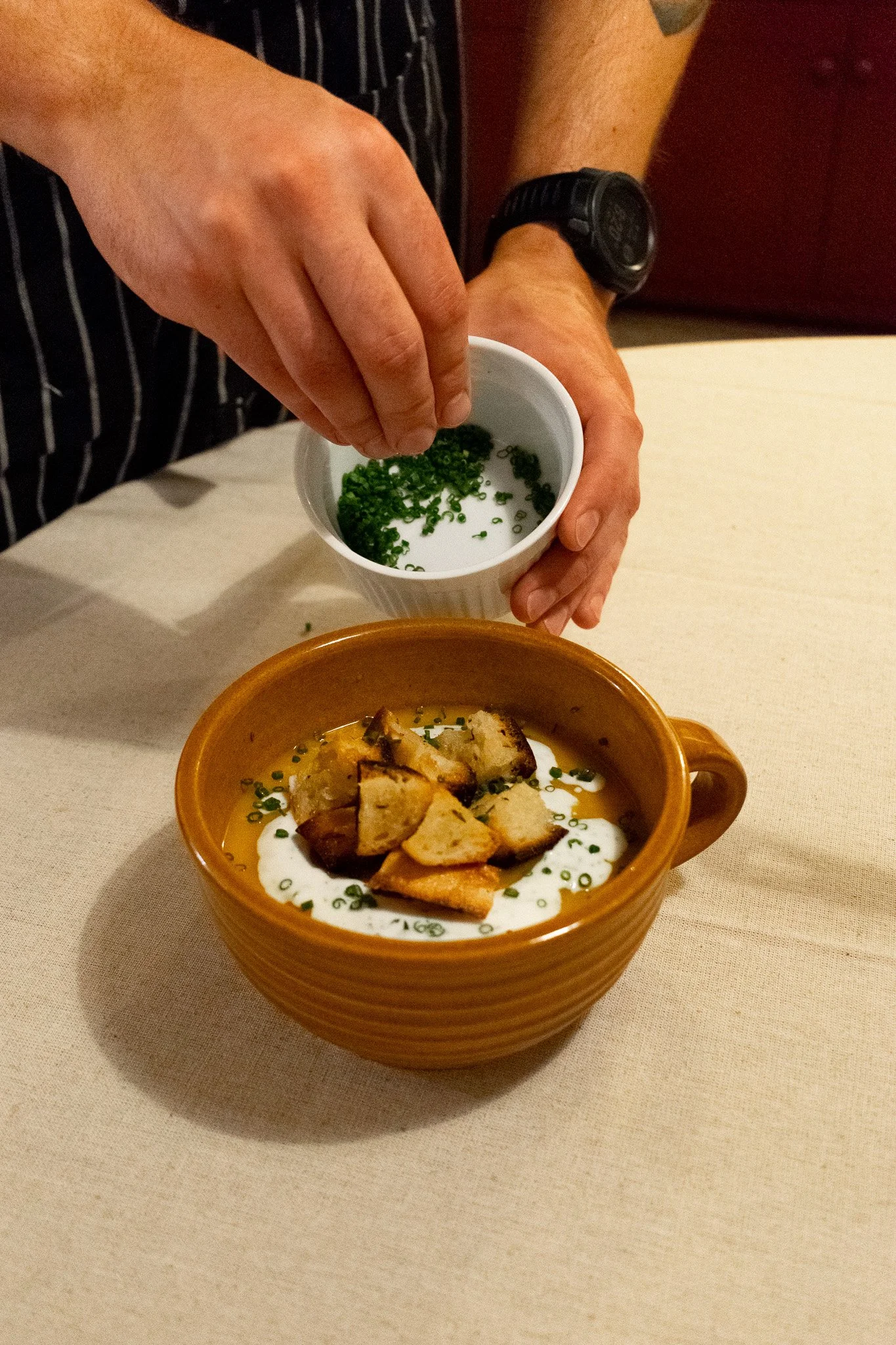A person adding chopped green herbs to a bowl of creamy soup topped with croutons, on a beige table.