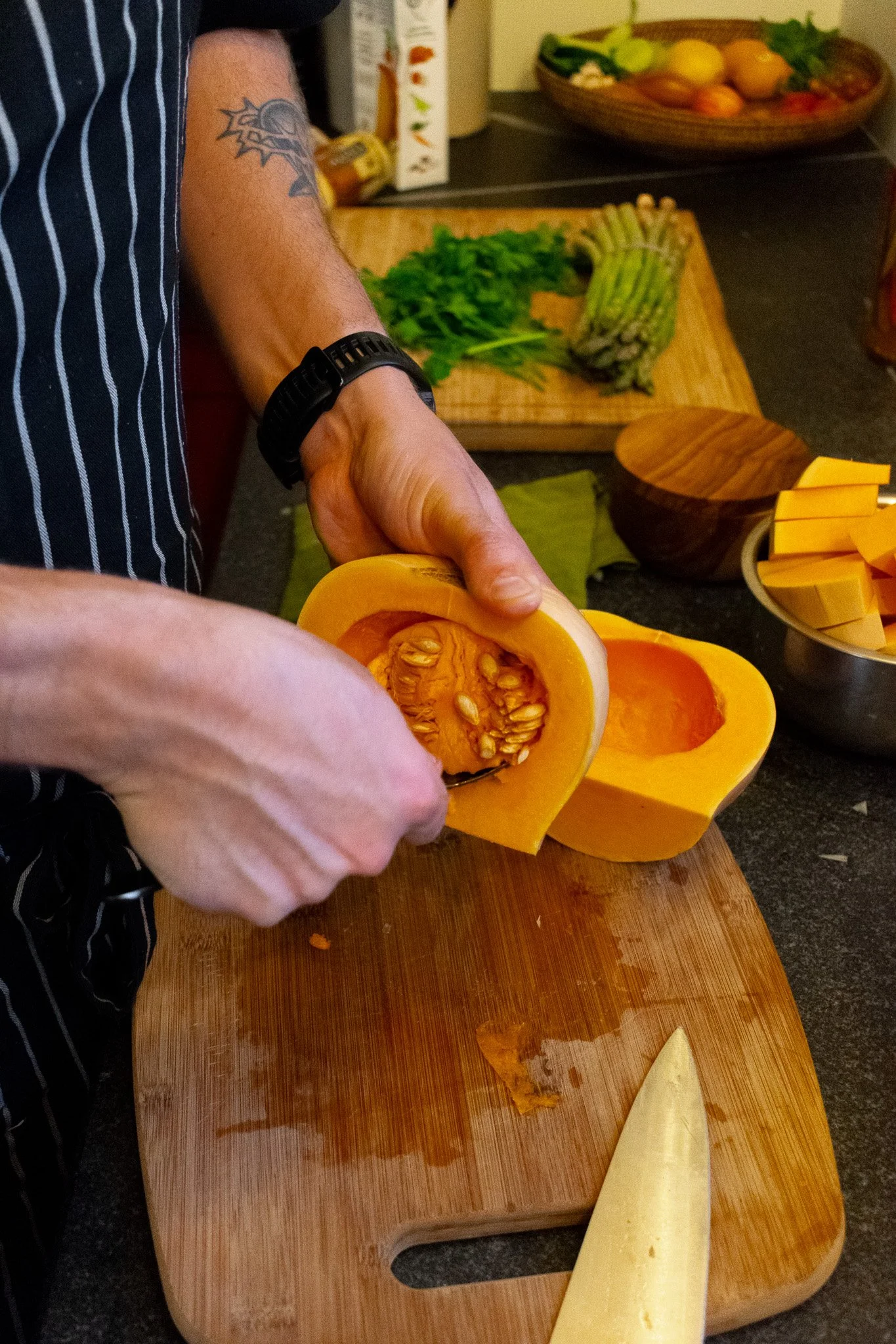 Person slicing a yellow squash on a wooden cutting board, with other vegetables and kitchen items in the background.