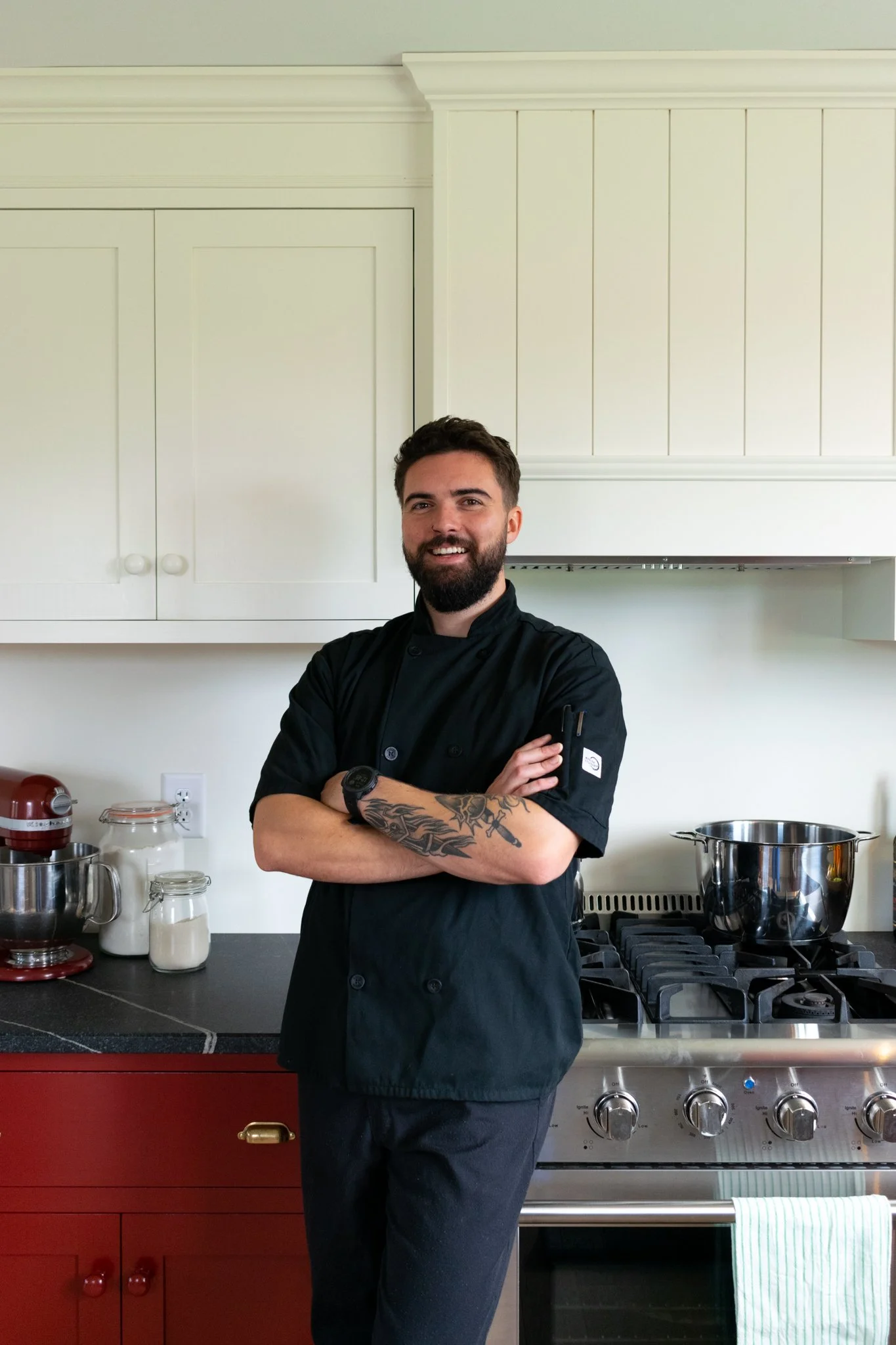 A male chef with a beard, wearing a black chef's coat, standing in a kitchen with arms crossed, smiling at the camera.