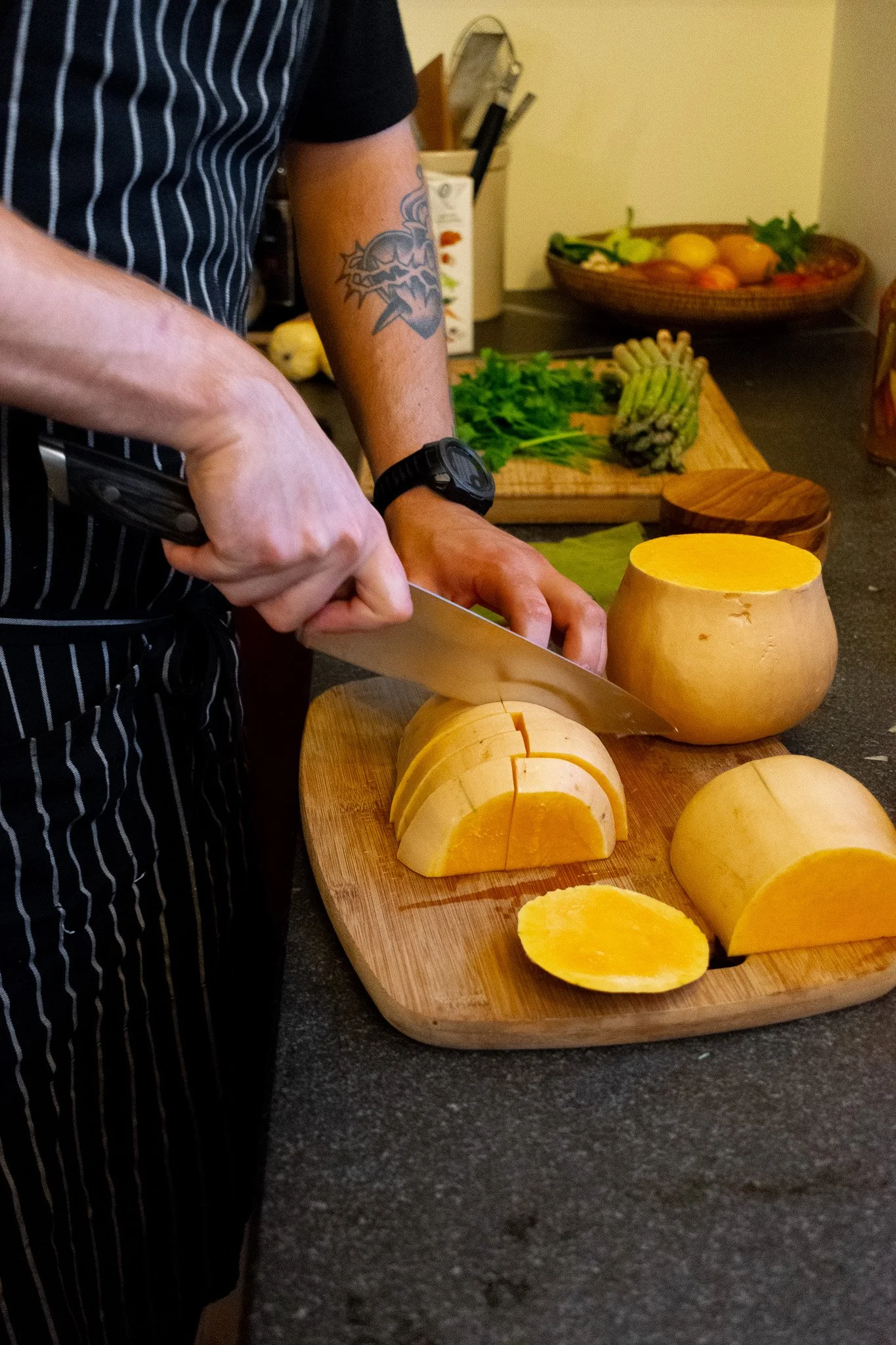 Chef slicing a large yellow pumpkin on a wooden cutting board in a kitchen, with various vegetables including asparagus, lettuce, and tomatoes in the background.