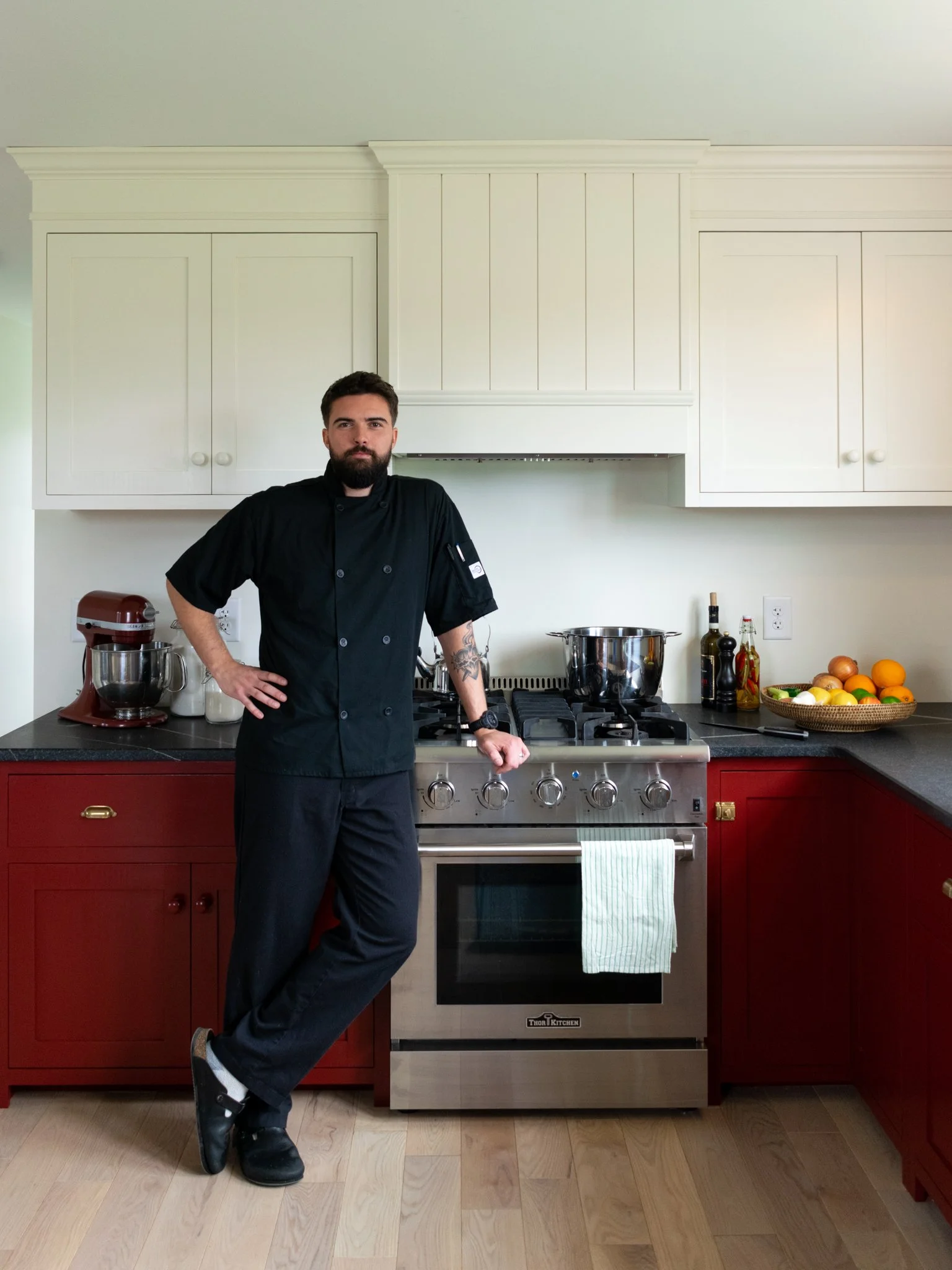 Chef dressed in black chef's uniform, standing in a kitchen with red and white cabinets, a stove, and various kitchen items including a stand mixer, fruits, and bottles.