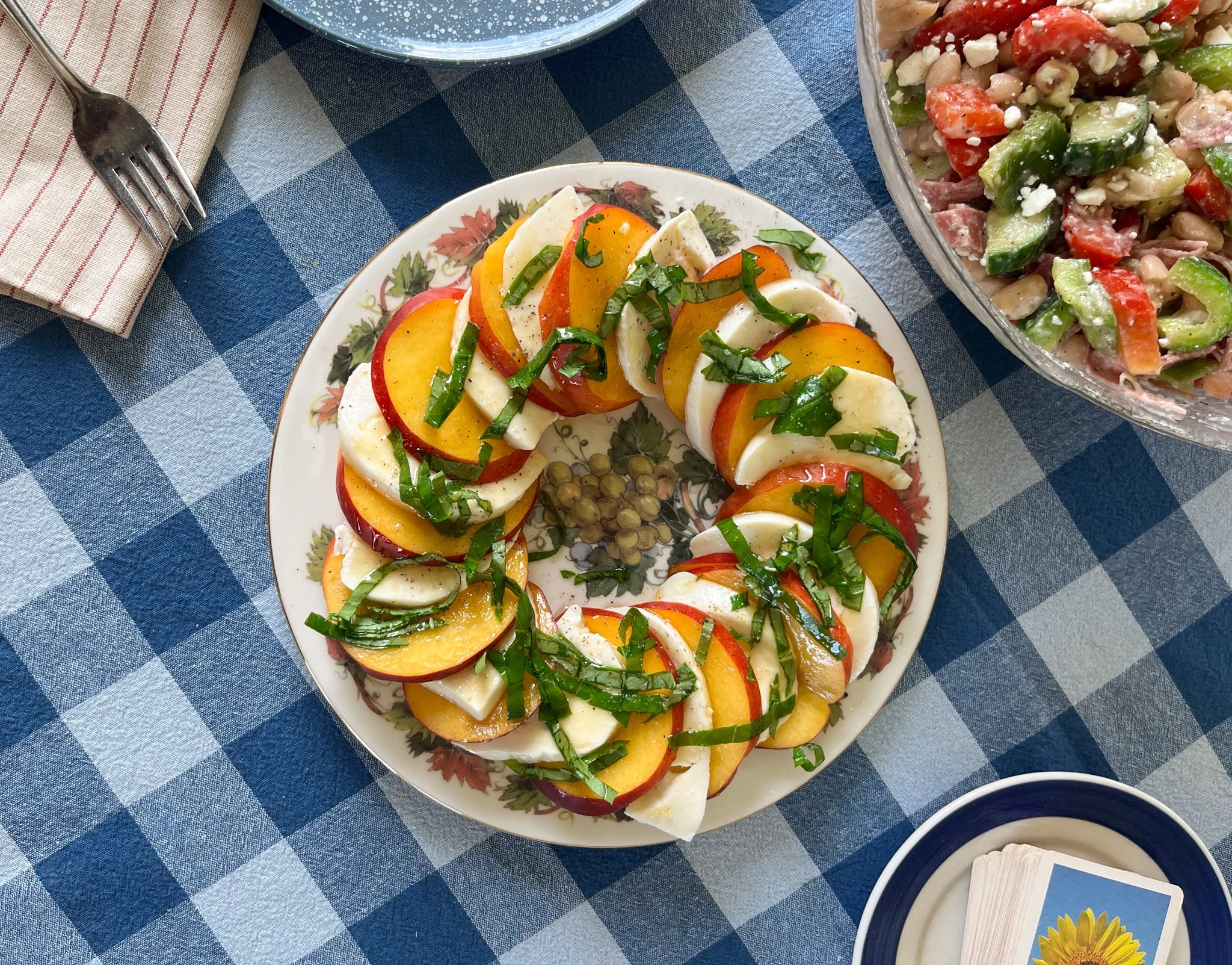 A plate of Caprese salad with sliced tomatoes, mozzarella cheese, basil, and a drizzle of olive oil, garnished with chopped green herbs, on a blue and white checkered tablecloth.
