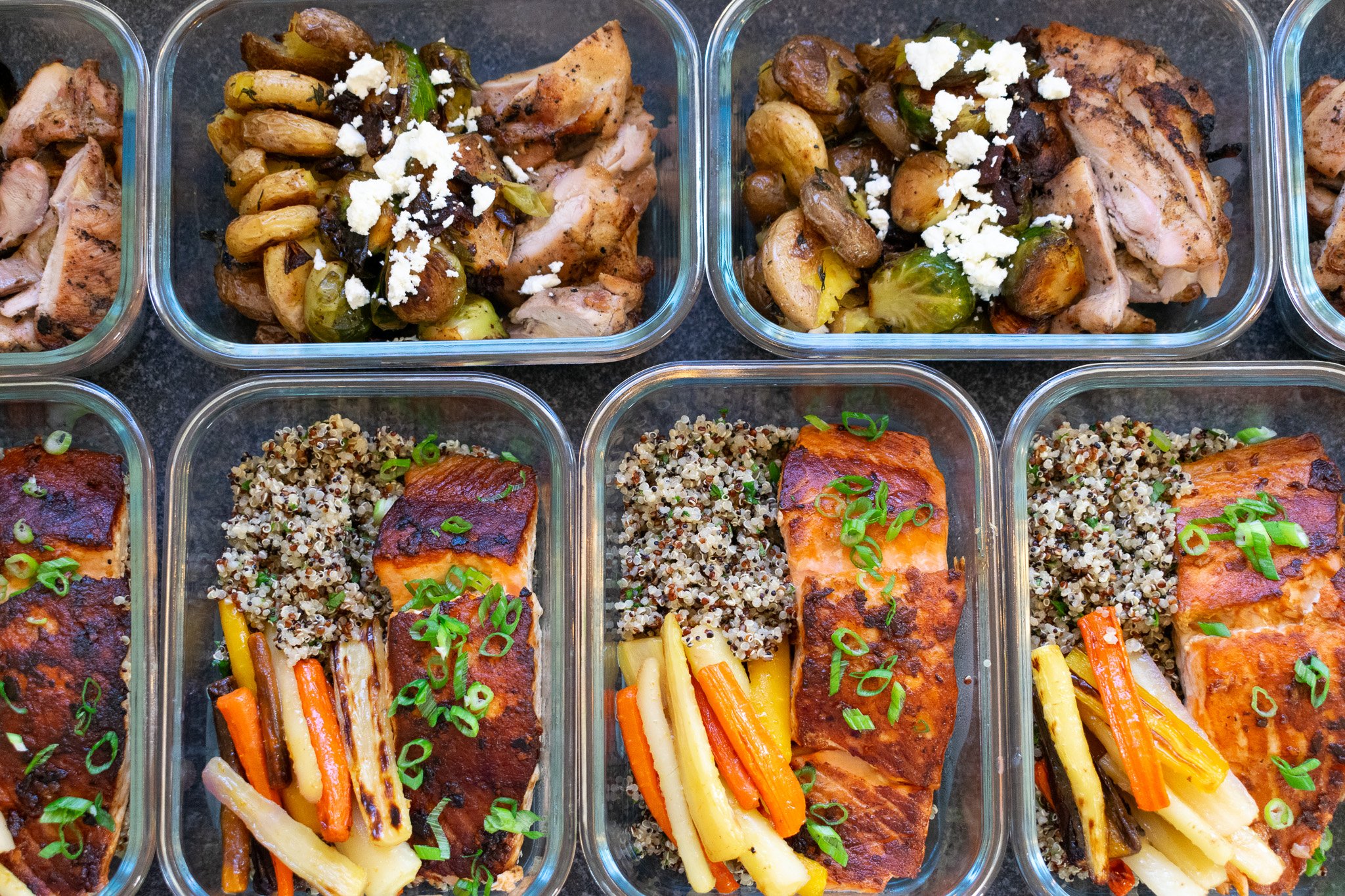 Multiple glass meal prep containers filled with cooked food including grilled chicken topped with vegetables and feta, and salmon fillets with quinoa and green onions.