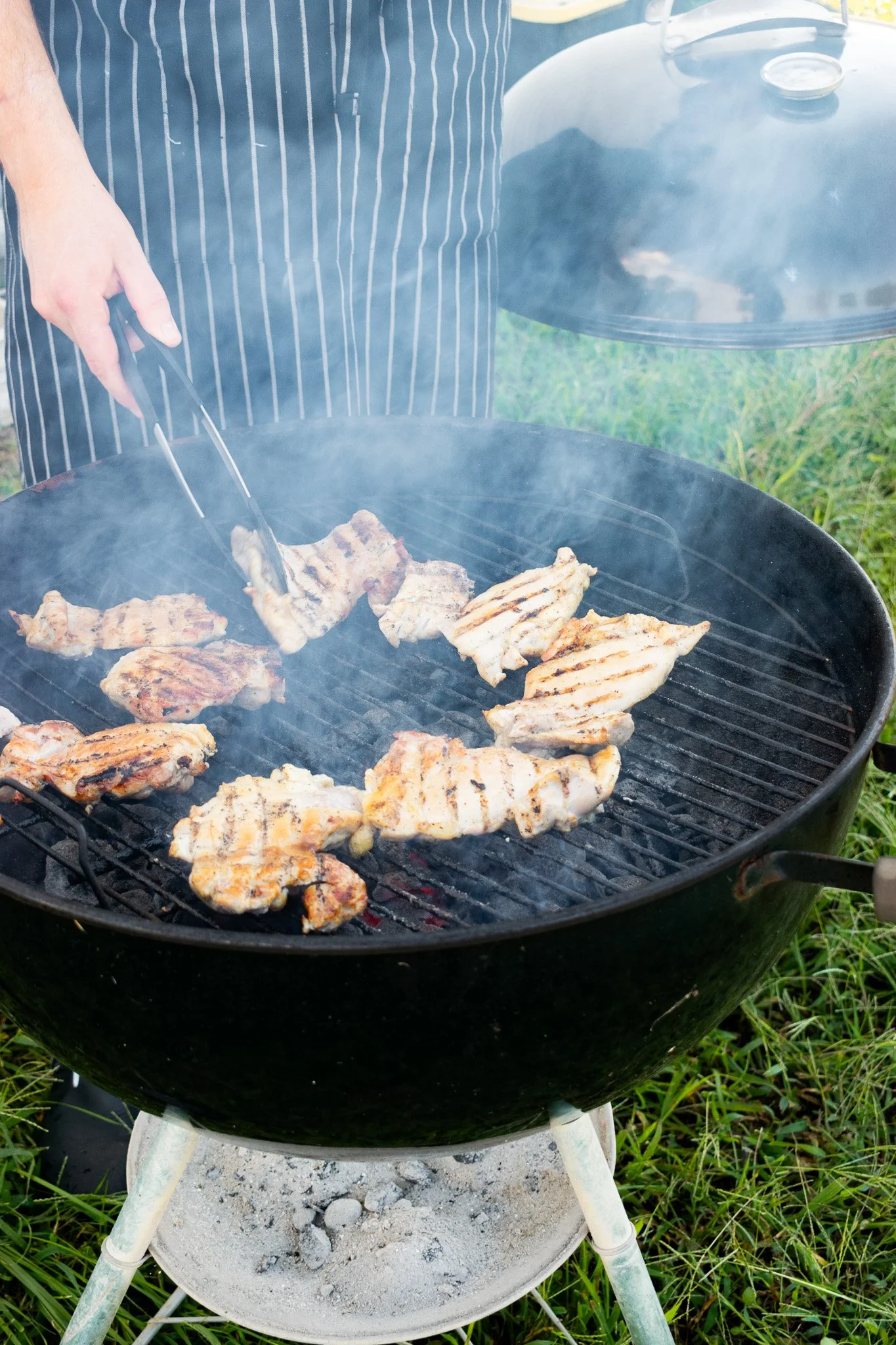 A barbecue grill outdoors with grilled chicken pieces cooking, covered in smoke. A person in a striped apron is using tongs to turn the chicken. The grill is set on a stand with ash and charcoal underneath, surrounded by grass.