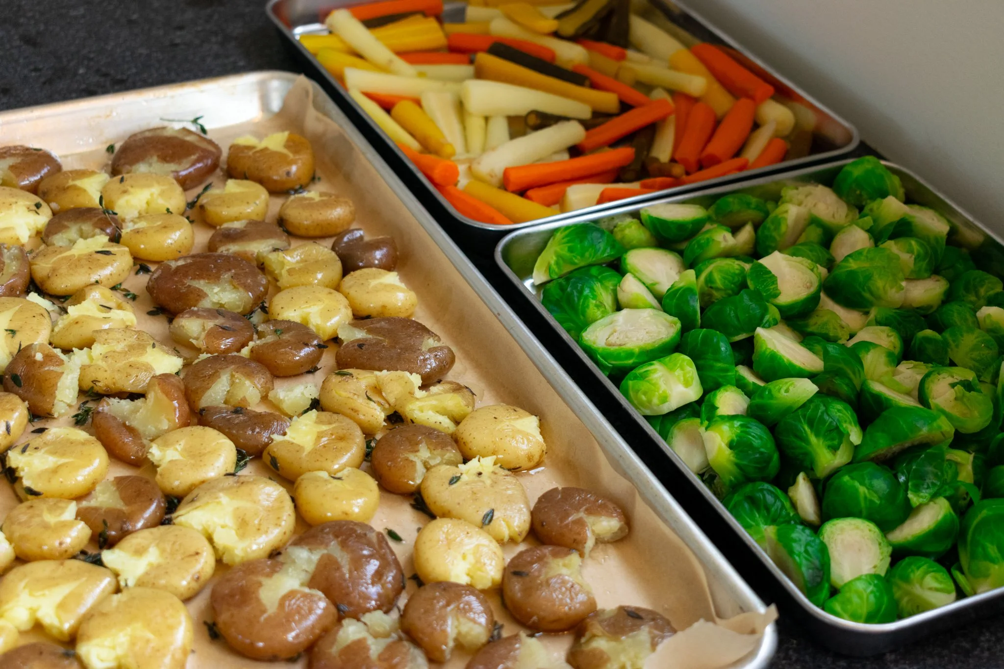Tray of roasted potatoes with herbs, containers of mixed roasted vegetables including carrots, parsnips, and Brussels sprouts on a black countertop.