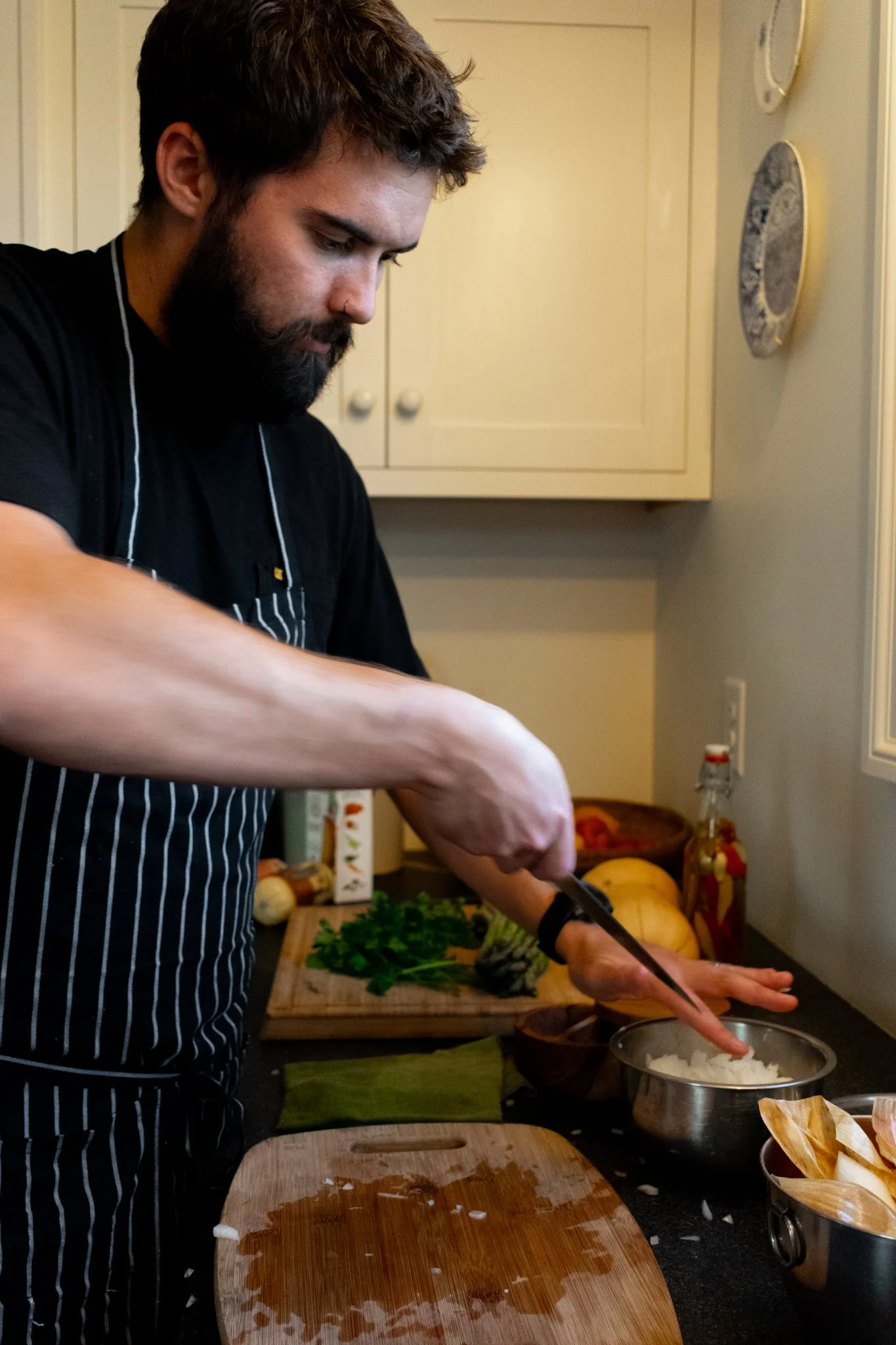 Chef wearing a black apron with white stripes is chopping onions in a kitchen. There are various vegetables and kitchen items on the counter behind him.