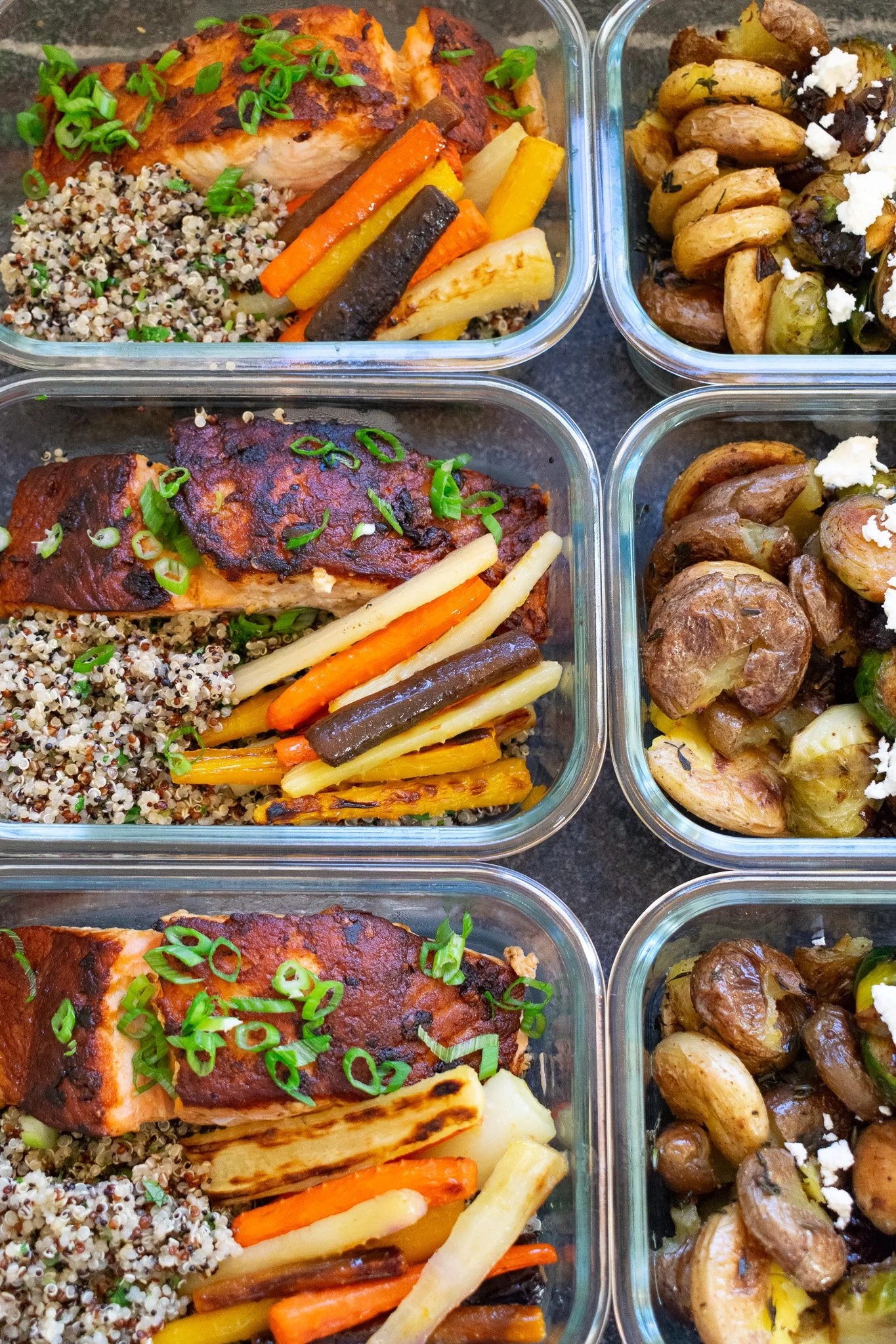 Glass meal prep containers filled with cooked salmon topped with chopped green onions, cooked quinoa, roasted mixed vegetables including carrots, parsnips, and sweet potatoes, and roasted potatoes with sautéed mushrooms, garnished with feta cheese.
