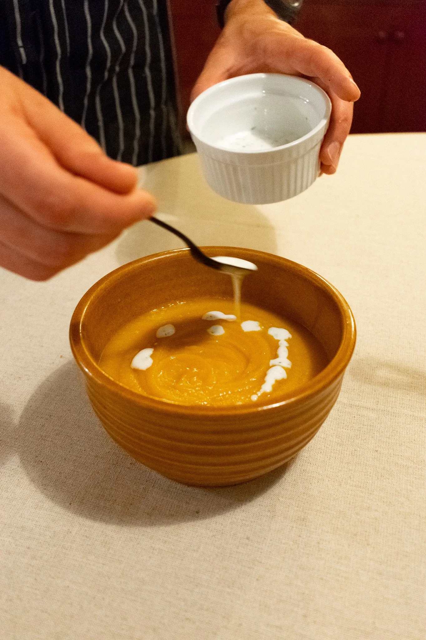 A person is pouring cream or milk from a small white ramekin into a bowl of pumpkin or squash soup, which is garnished with a swirl of cream.