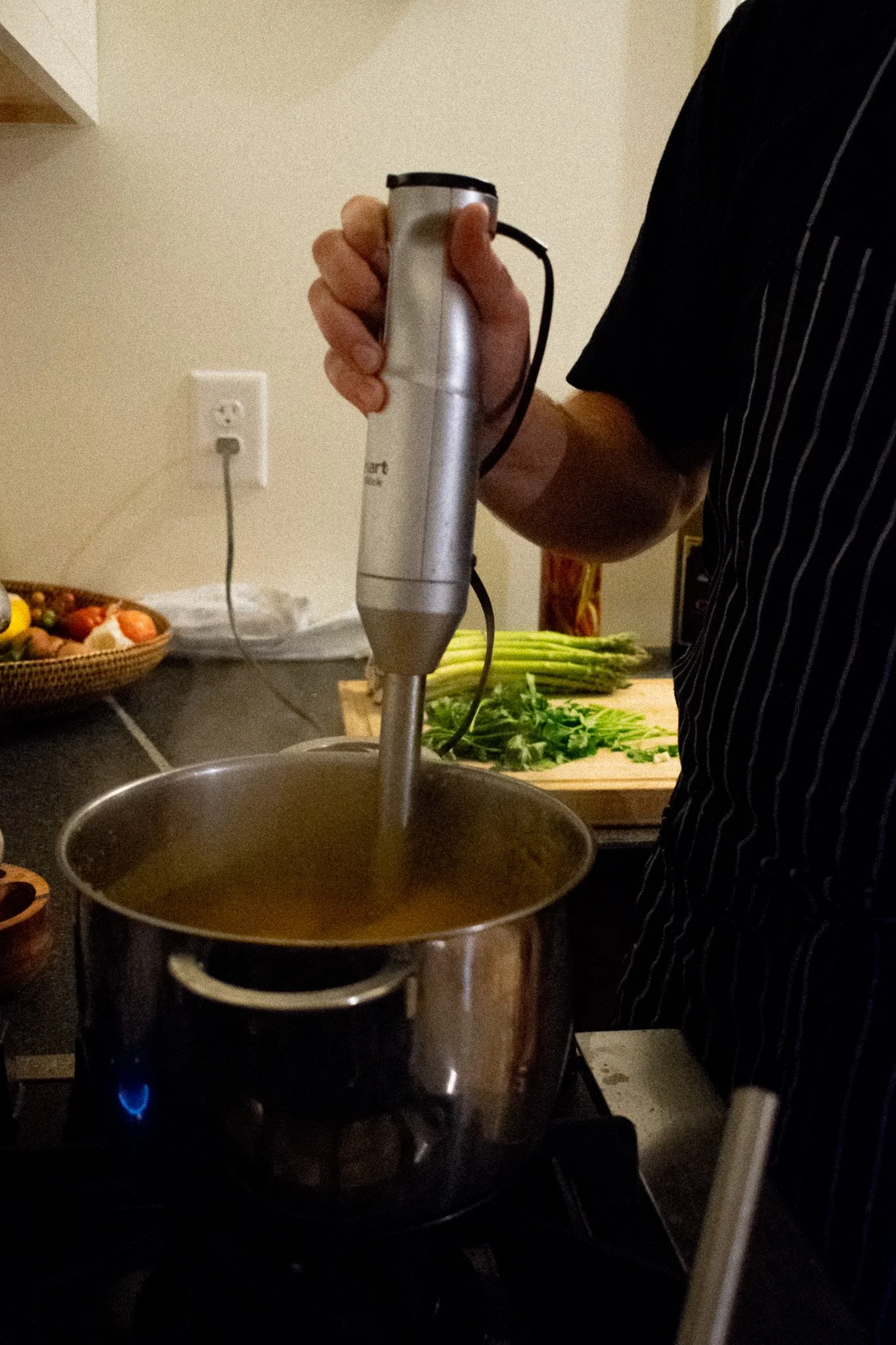 Chef cooking on stovetop with a pot and a hand-held immersion blender blending soup, vegetables on cutting board in the background.