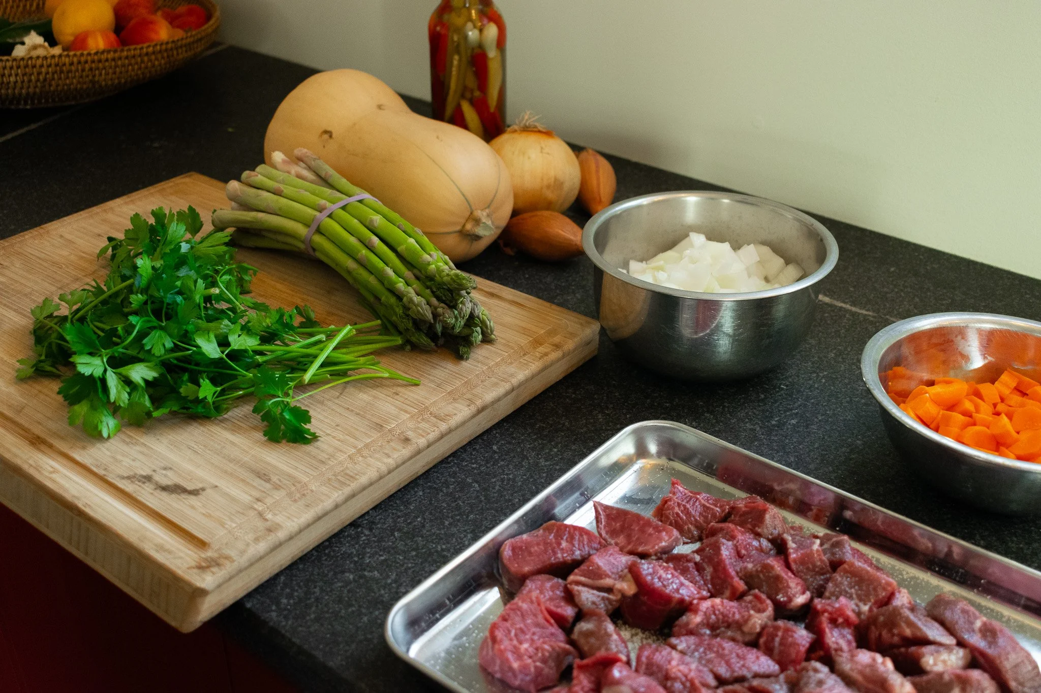 A kitchen countertop with fresh herbs, chopped onions in a metal bowl, sliced carrots, and raw beef on a tray, with onions and a basket of other fruits in the background.