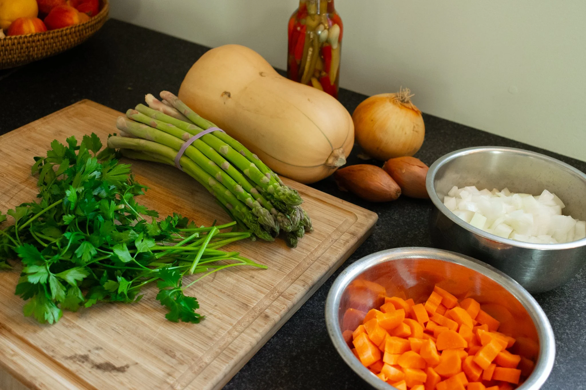 Fresh vegetables including parsley, asparagus, butternut squash, onions, garlic cloves, chopped onions, chopped carrots on kitchen counter and a string jar of pickled vegetables in the background.