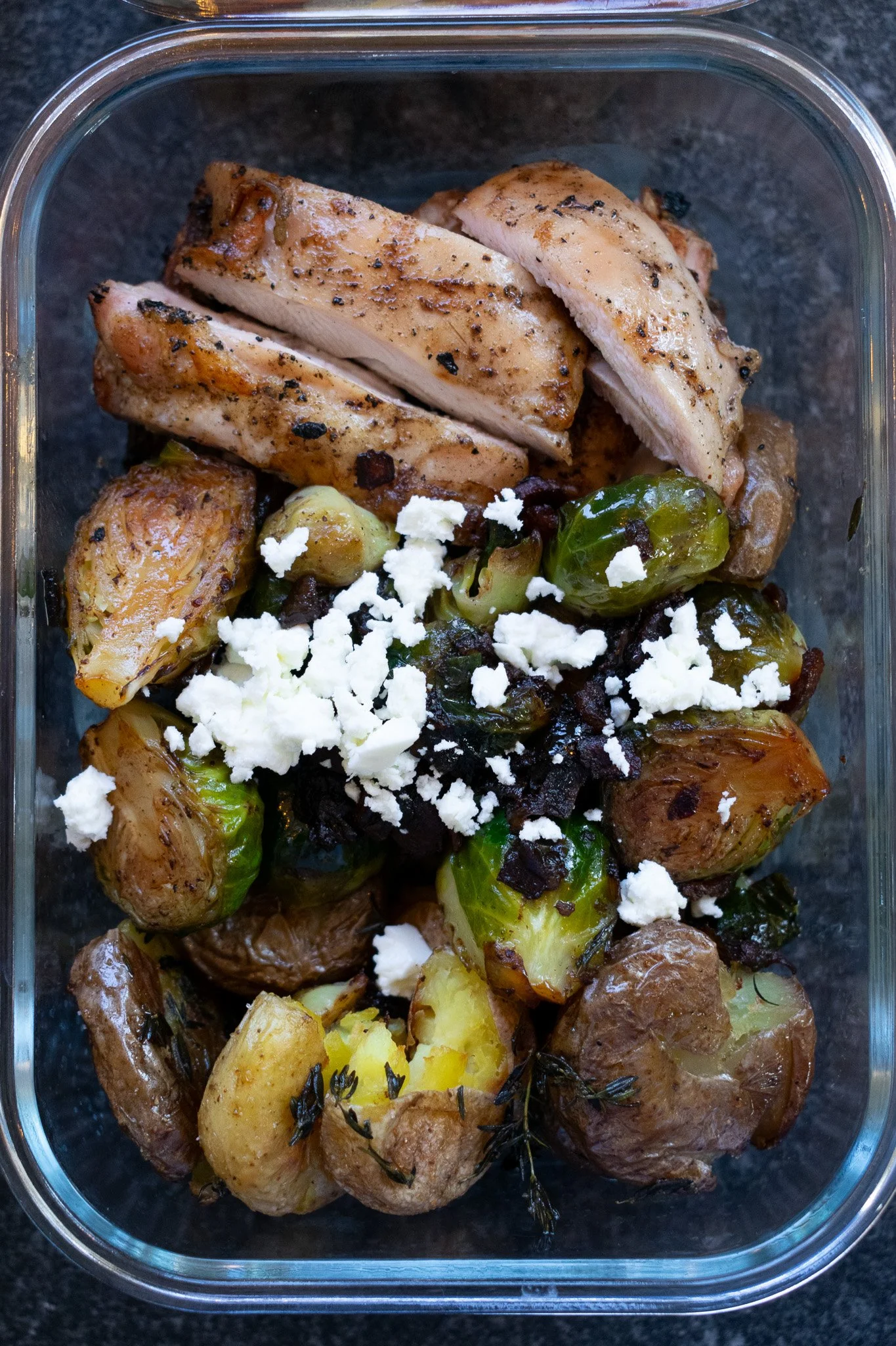 Cooked chicken breasts with seasoning, grilled Brussels sprouts, roasted potatoes, blue cheese crumbles, and sprigs of thyme in a glass container.