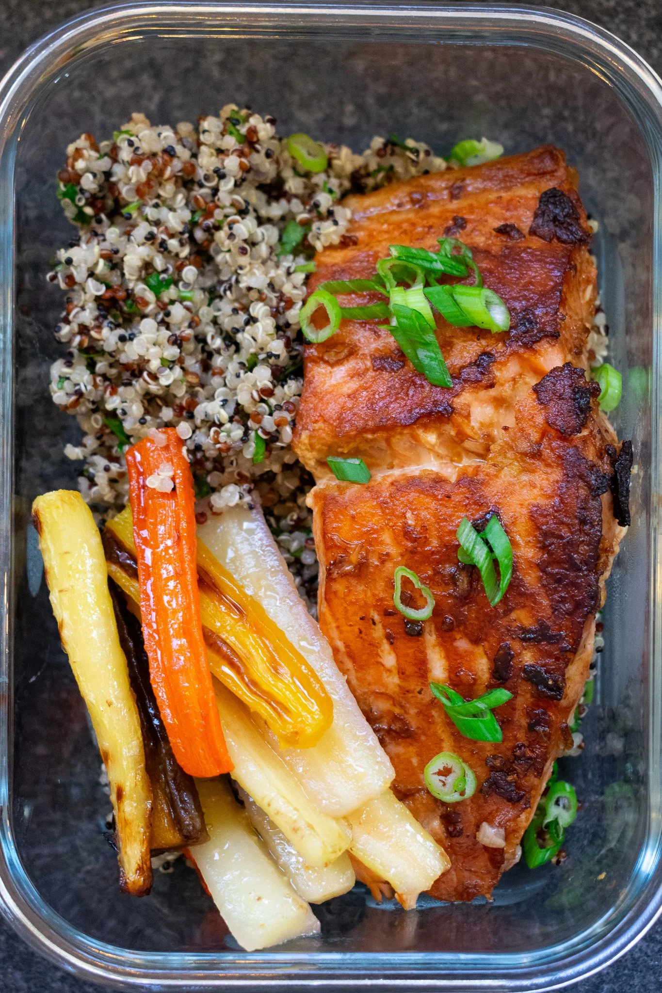 Baked salmon fillet garnished with chopped green onions, served with roasted assorted vegetables including carrots, yellow squash, and eggplant, alongside cooked quinoa in a glass container.