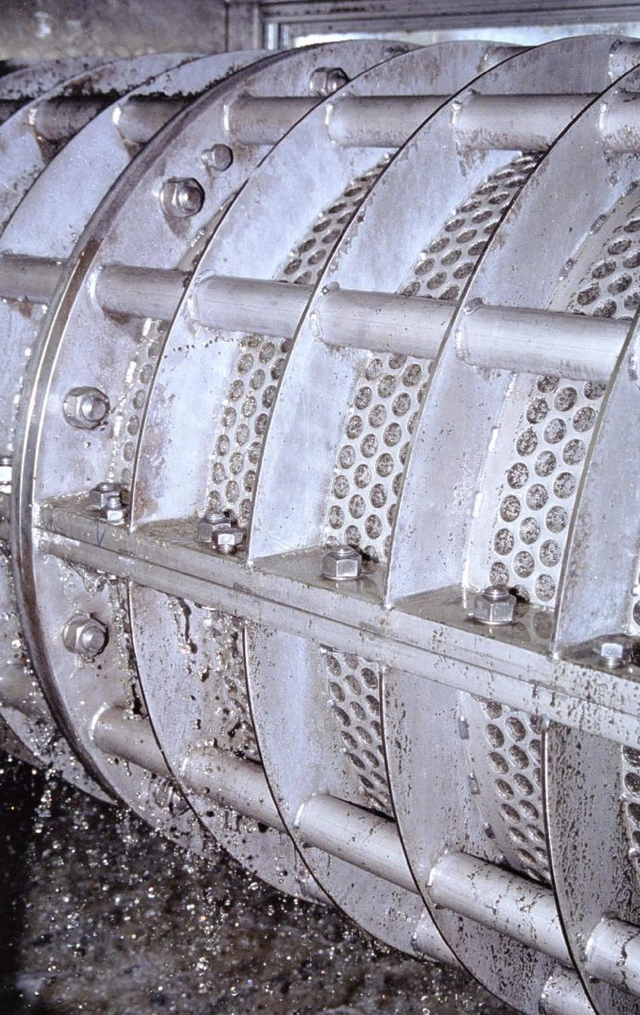 A close-up of a metallic industrial machine part with tubes, bolts, and perforated metal plates, appearing wet or dirty.