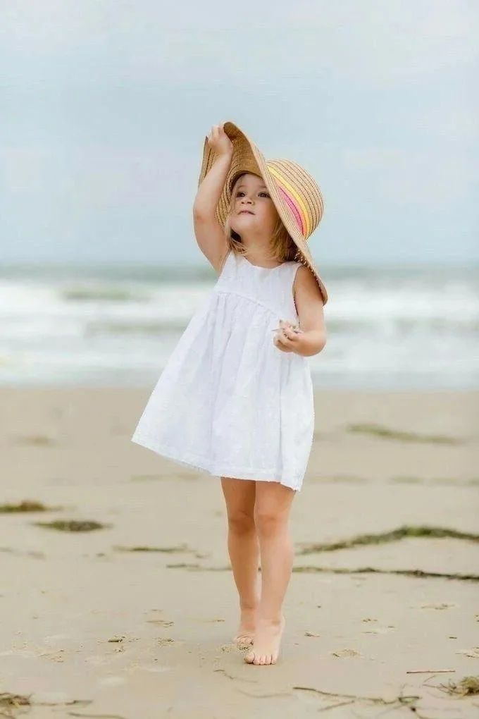 Une petite fille en robe blanche et chapeau de plage, marchant pieds nus sur la plage, avec la mer en arrière-plan.