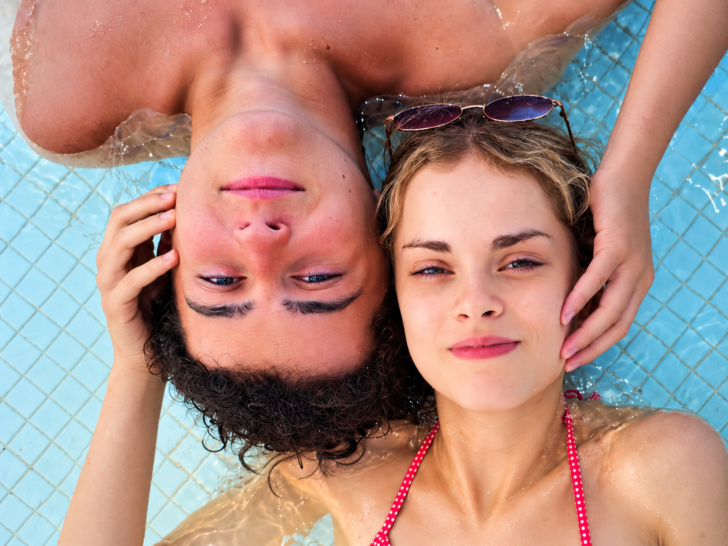 Deux jeunes femmes détendues flottant dans une piscine, tête contre tête, avec des sourires, une porte-sunglasses sur une, et une peau bronzée en maillot en polka dots rouge et blanc.