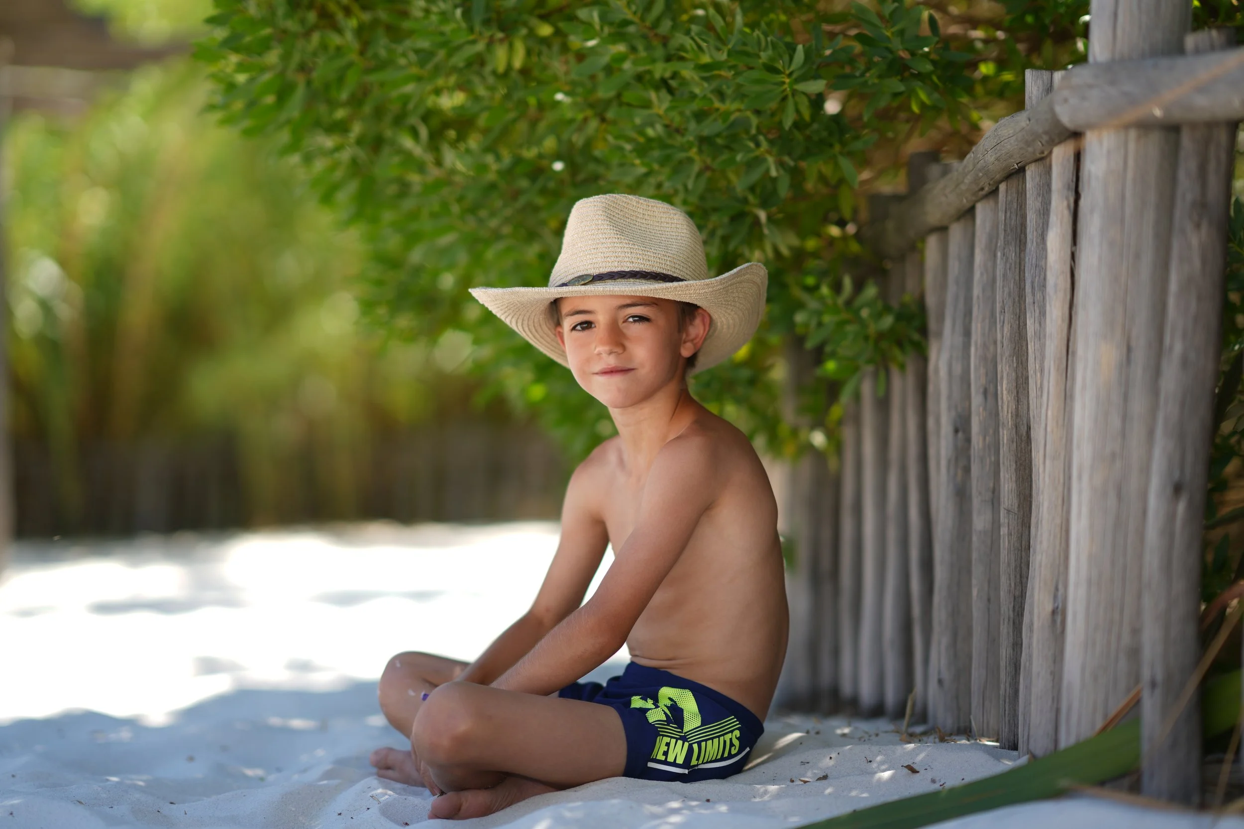Un jeune garçon est assis sur le sable, portant un chapeau de paille et un short de bain bleu avec des motifs verts, près d'une clôture en bois et de verdure, sous un ciel ensoleillé.