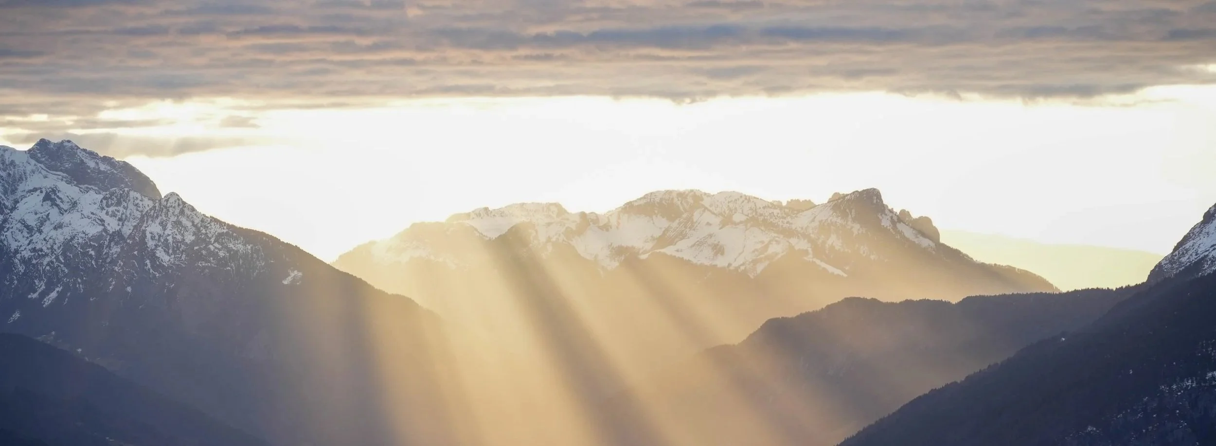 Montagnes enneigées sous un ciel nuageux avec des rayons de soleil brillants.