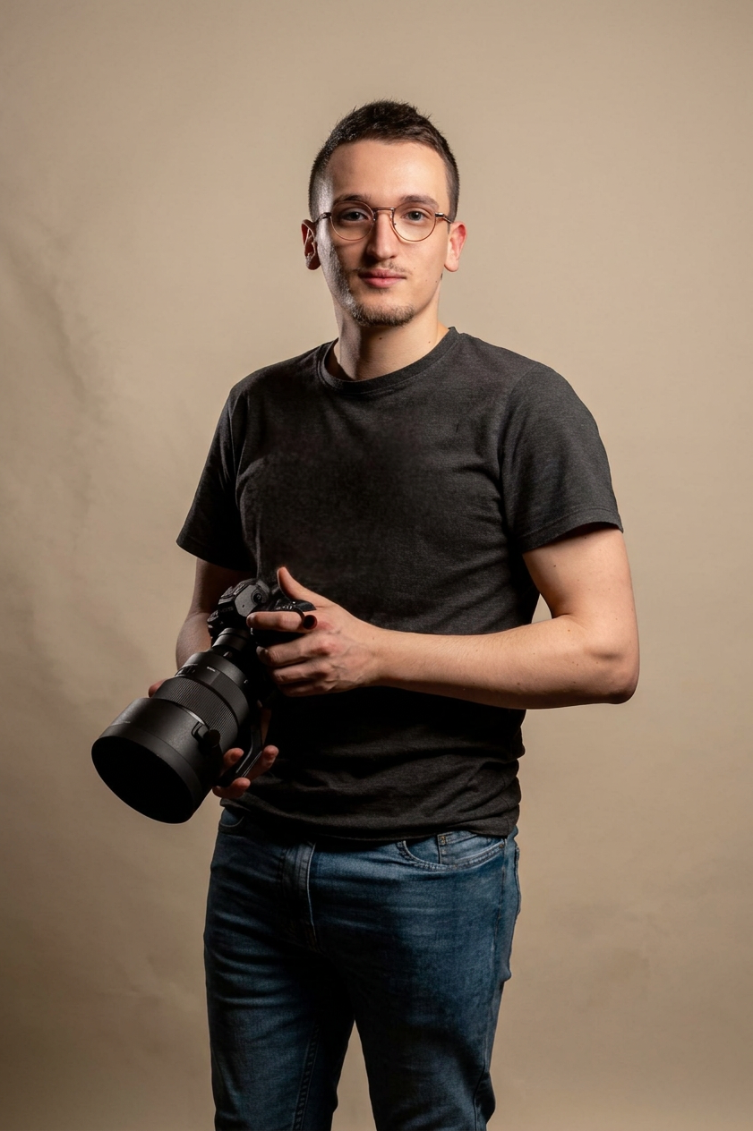 Jeune homme tenant un appareil photo réflex numérique, portant un t-shirt noir et des jeans, pose devant un fond beige.